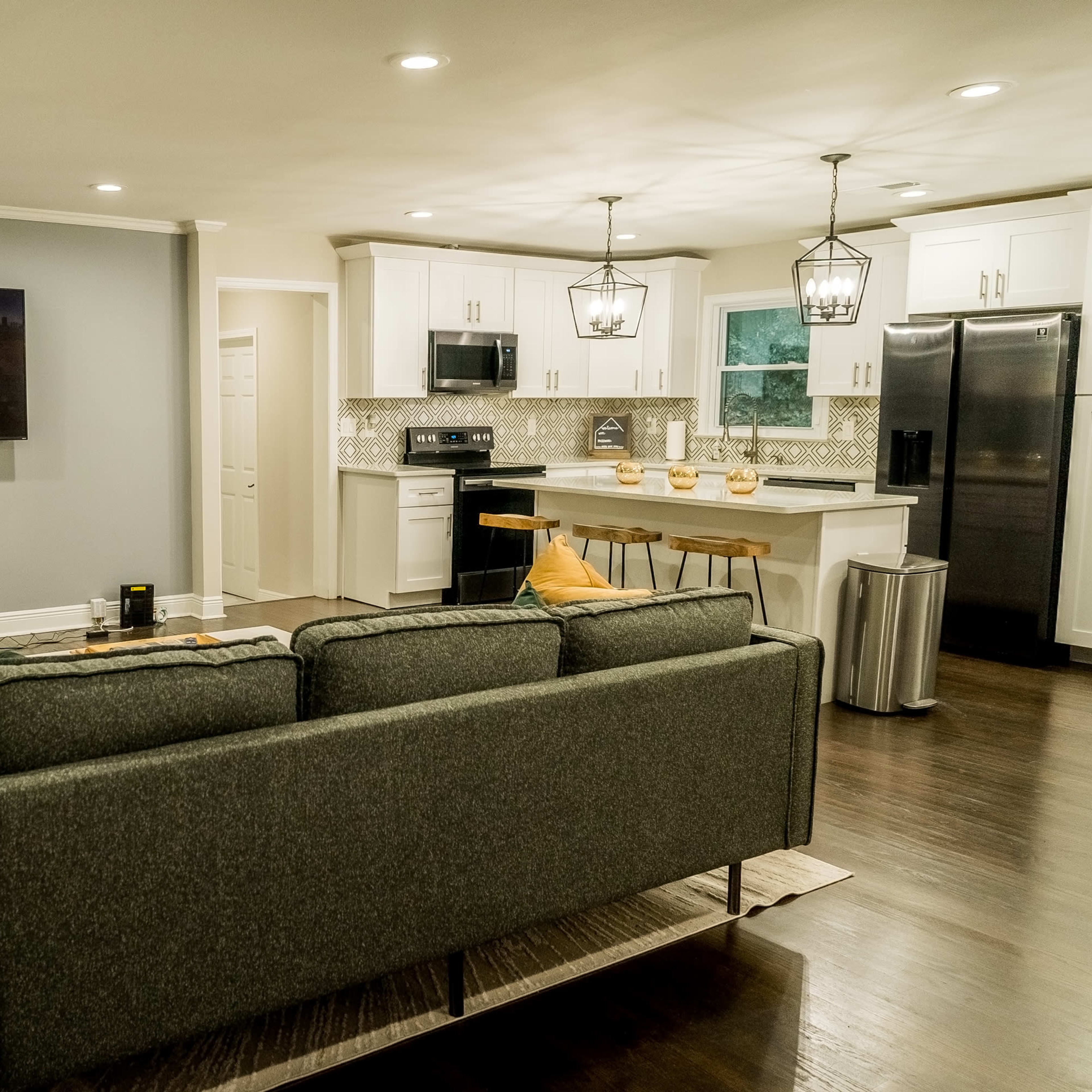 A modern living room that opens into a kitchen, featuring a gray sofa, a television on the wall, and white cabinetry.