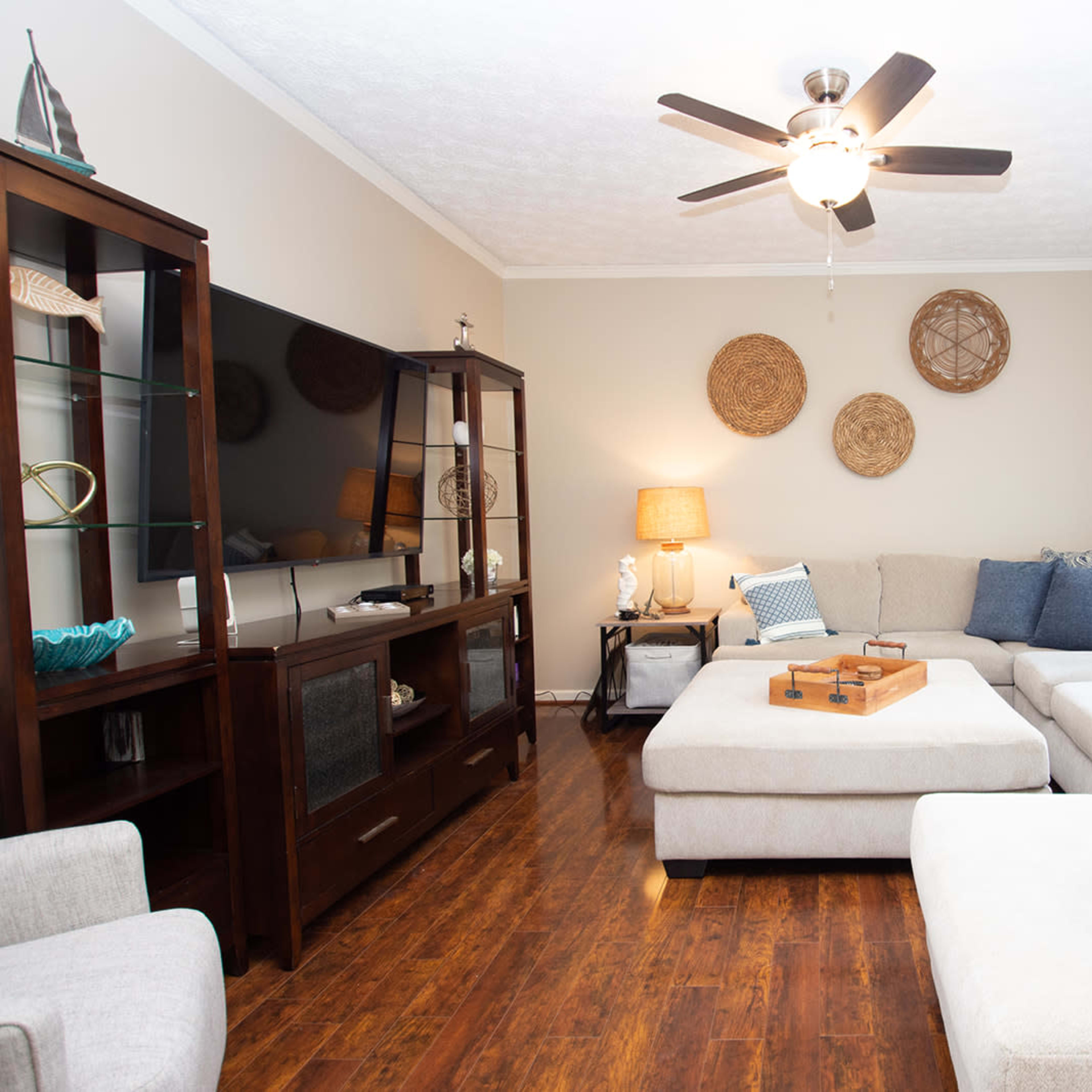 A large, light-colored sectional sofa is positioned in a cozy living room with hardwood floors, a flat-screen TV, and decorative wall hangings.