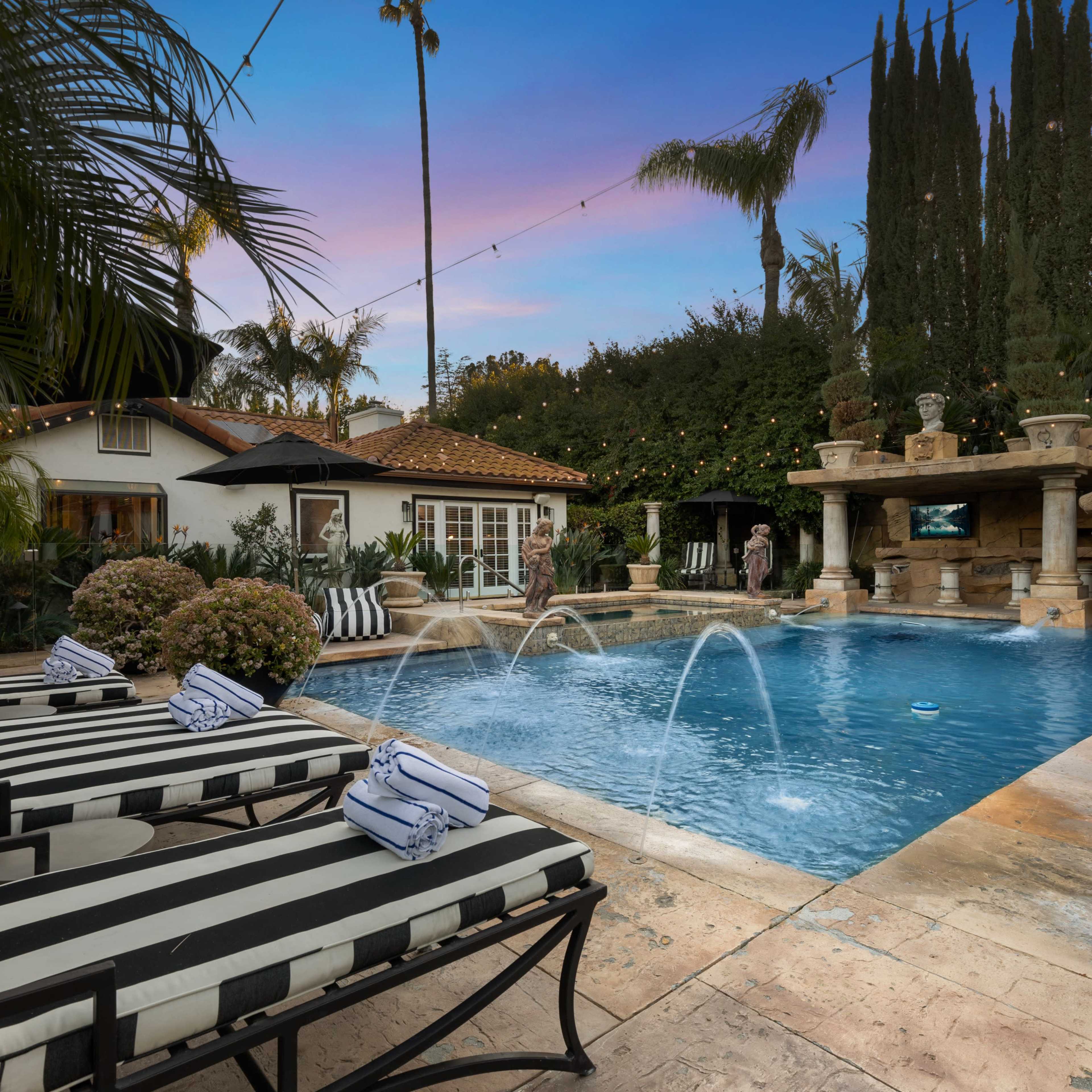 A pool with water features is surrounded by lounge chairs and lush landscaping, with a residential building and stone structure in the background.