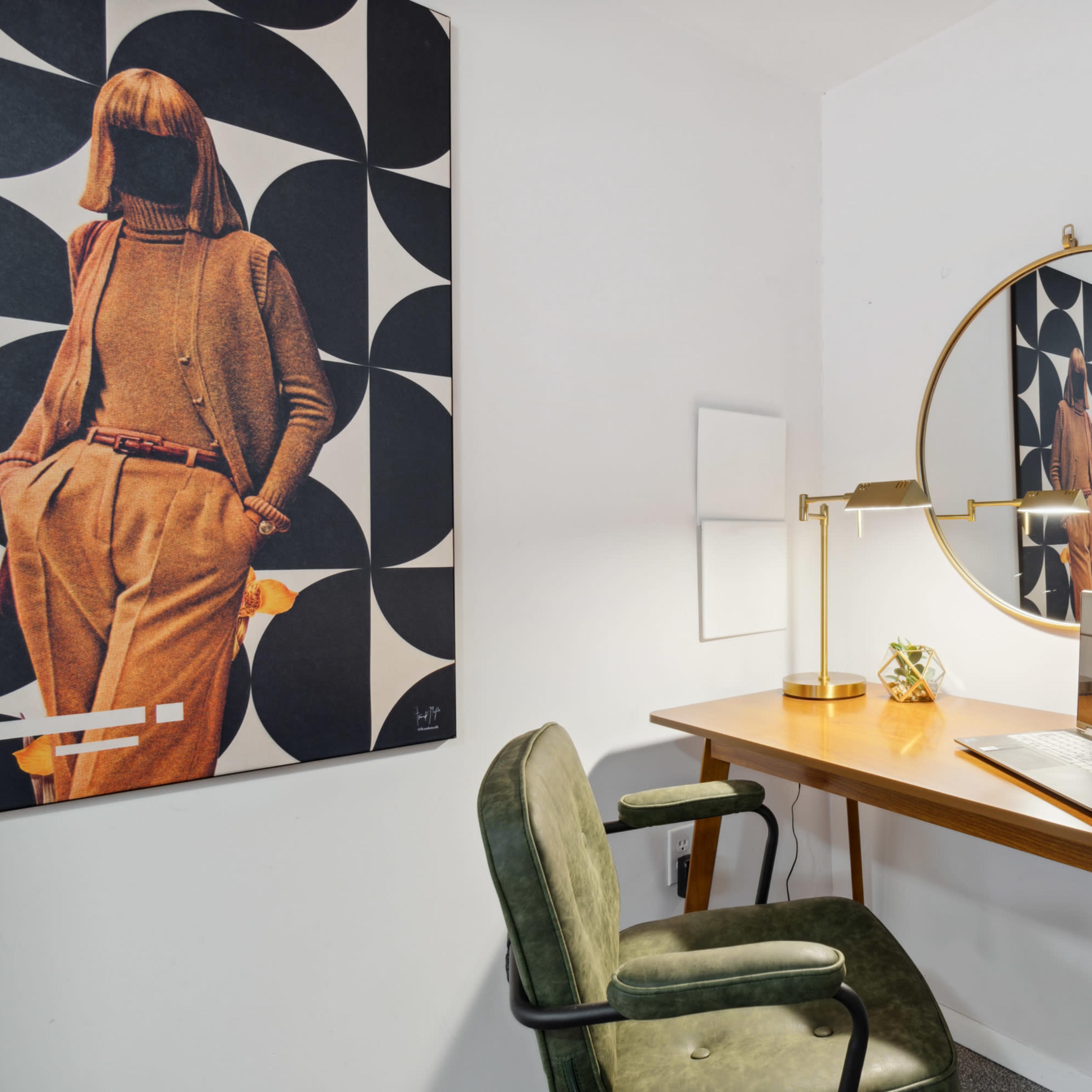 A desk with a laptop is positioned against a white wall, accompanied by a round mirror and a large framed artwork of a figure in a patterned background.