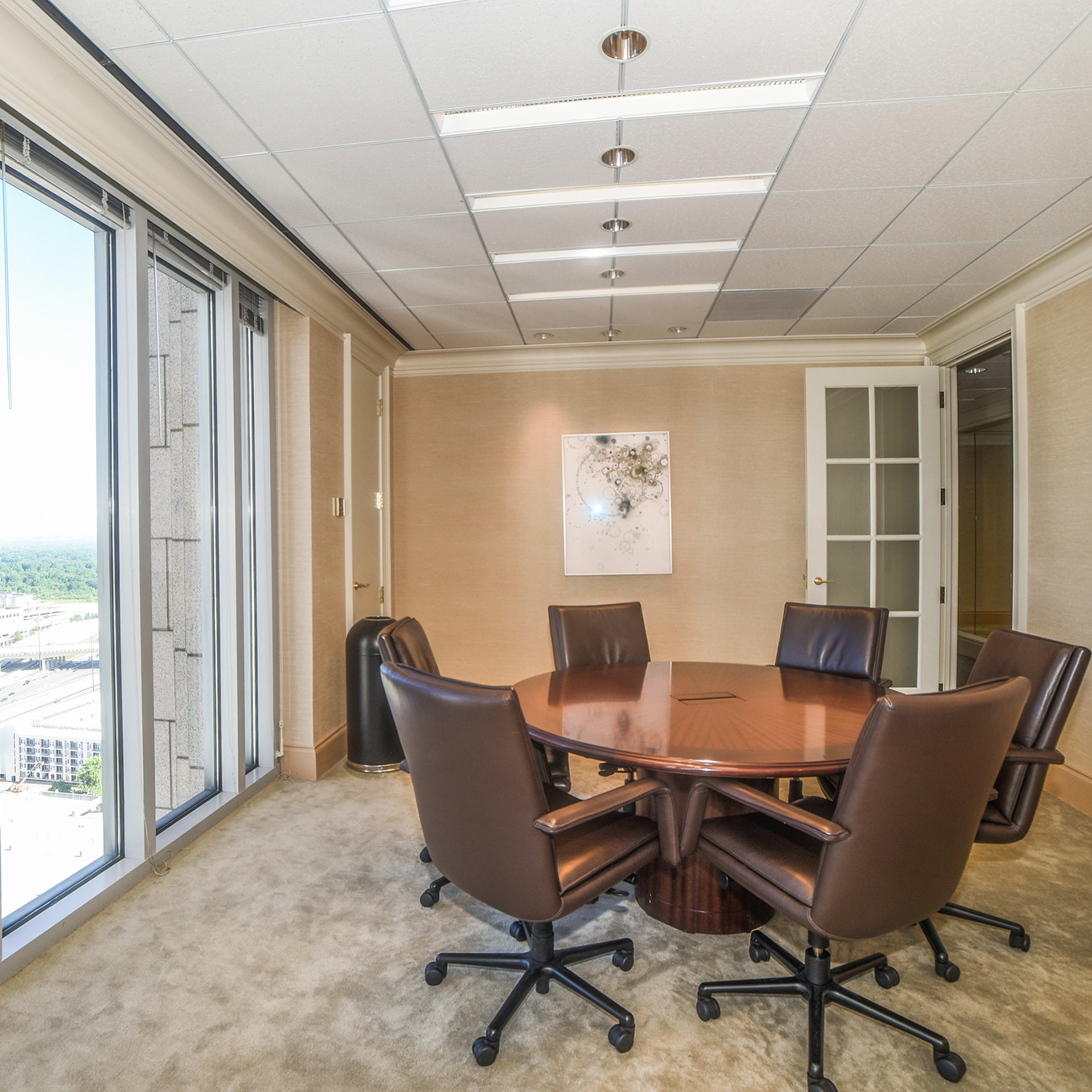 A conference room with a wooden round table surrounded by six brown chairs, featuring large windows that offer a view of the city.