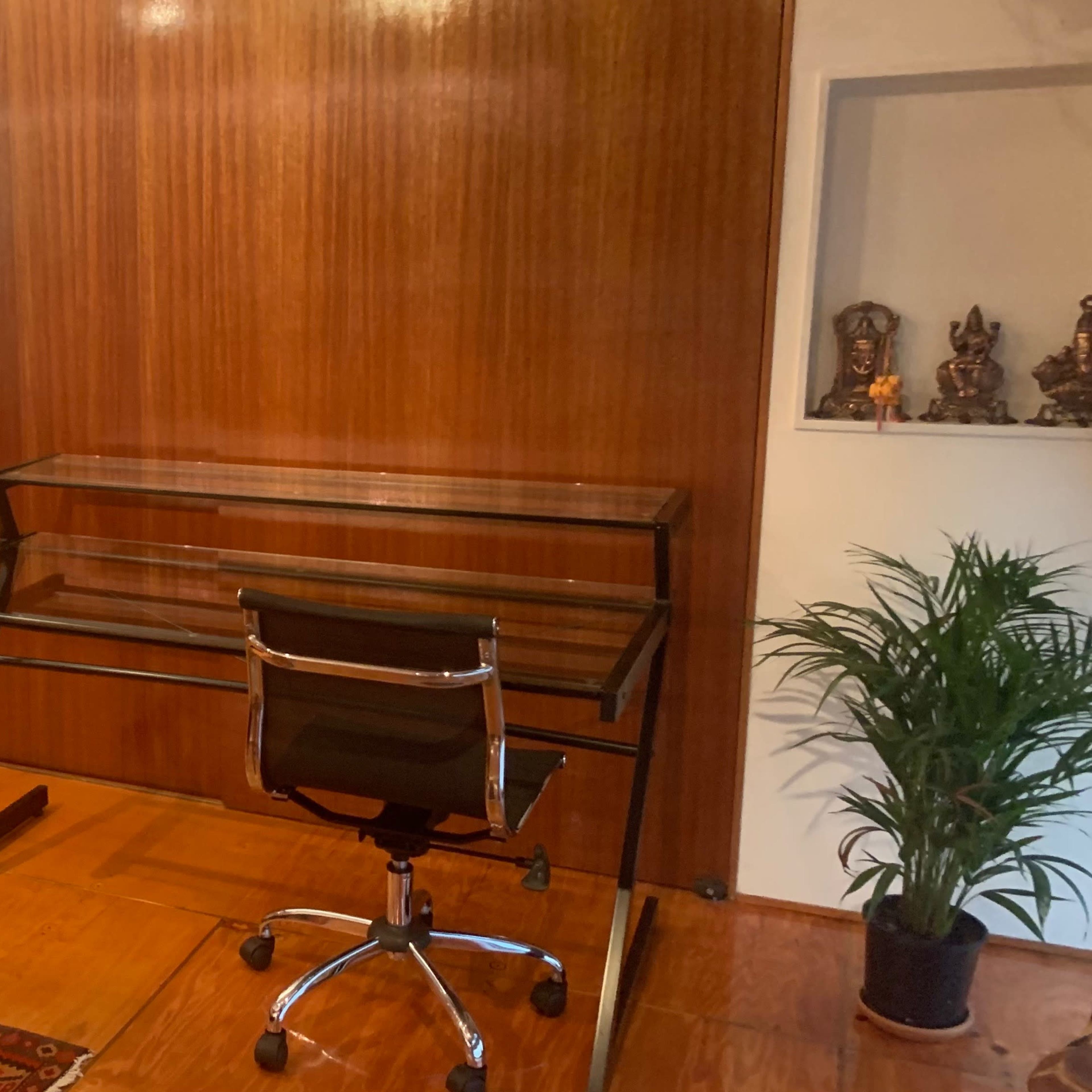 A minimalist office space featuring a glass desk with a rolling chair against a wooden wall, a television mounted on the wall, and decorative items on shelves.