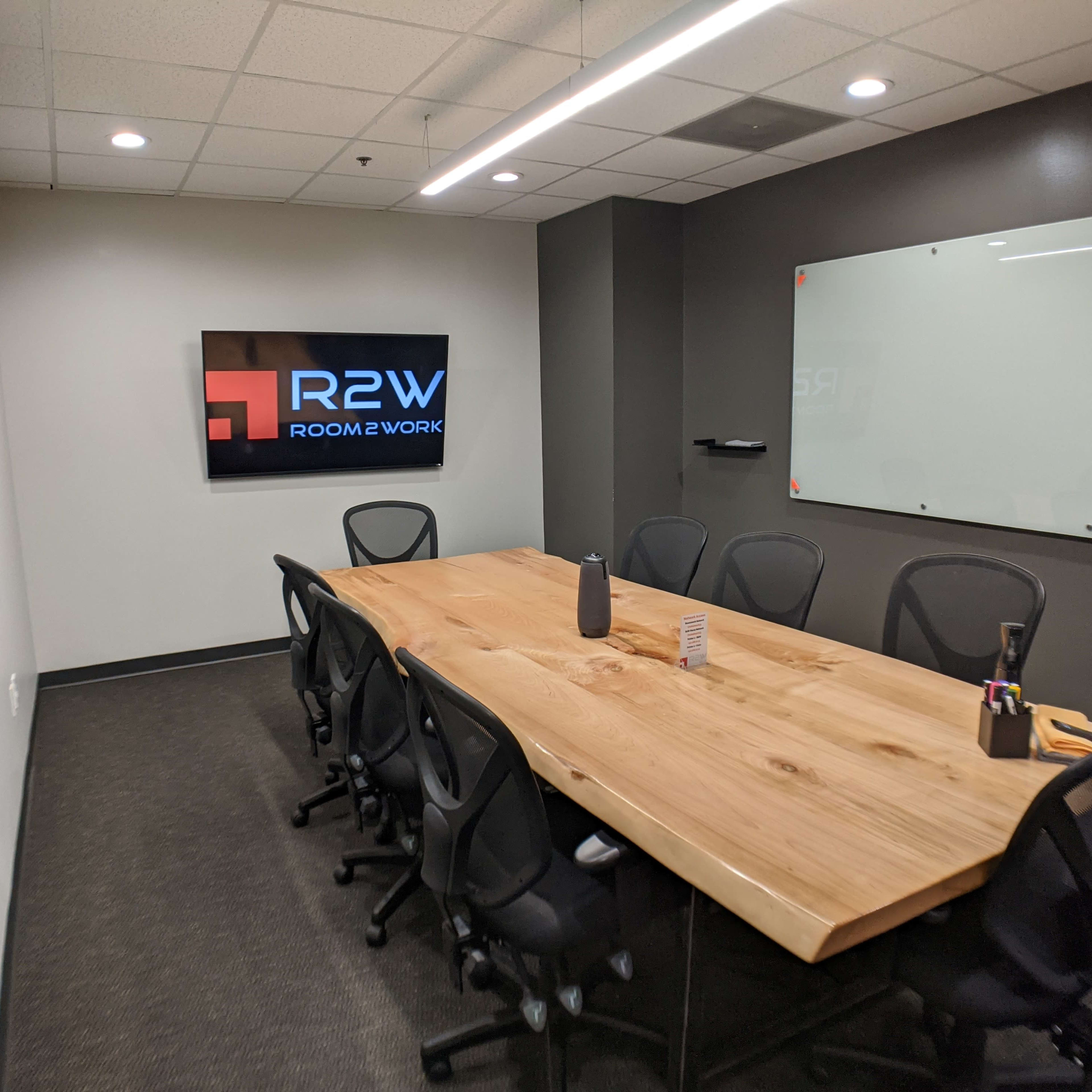 A modern conference room with a large wooden table surrounded by black chairs, featuring a wall-mounted screen displaying the "R2W Room 2 Work" logo.