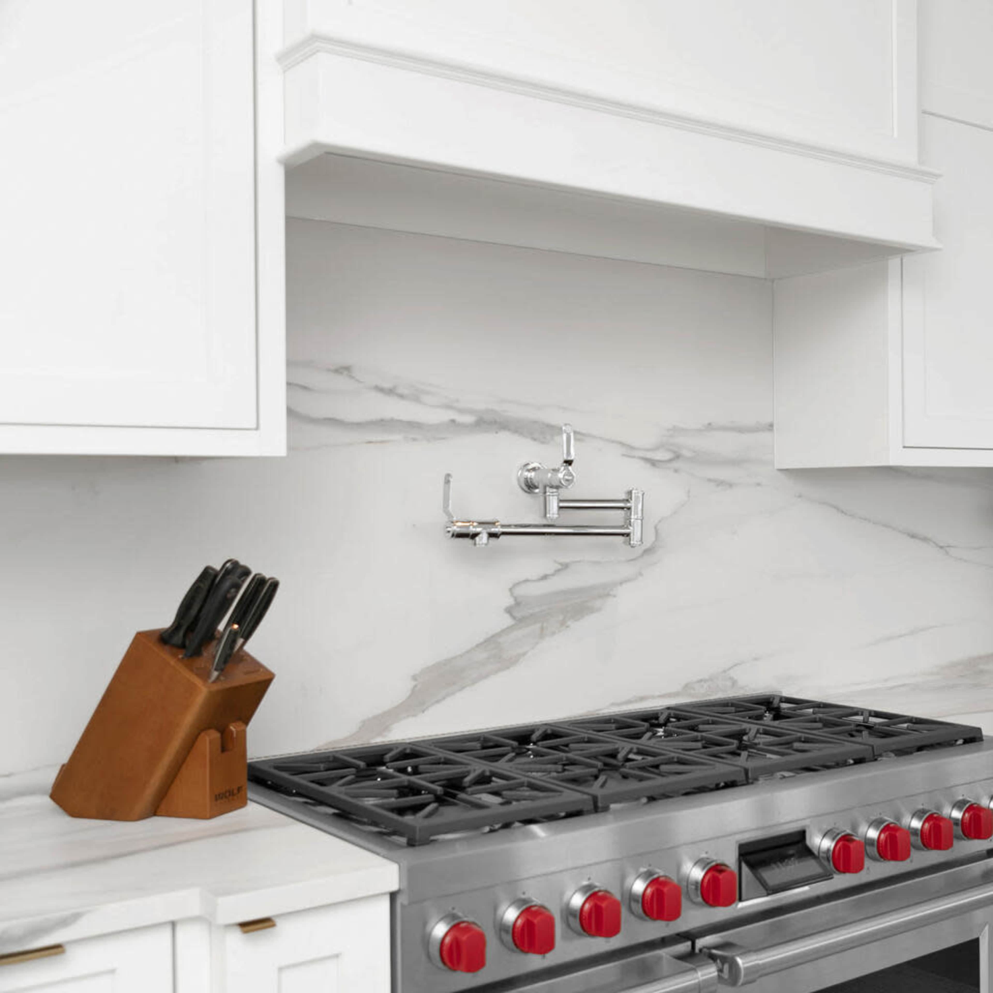 A modern kitchen features a stainless steel gas range with red knobs, a wall-mounted faucet, and white cabinetry against a marble backsplash.