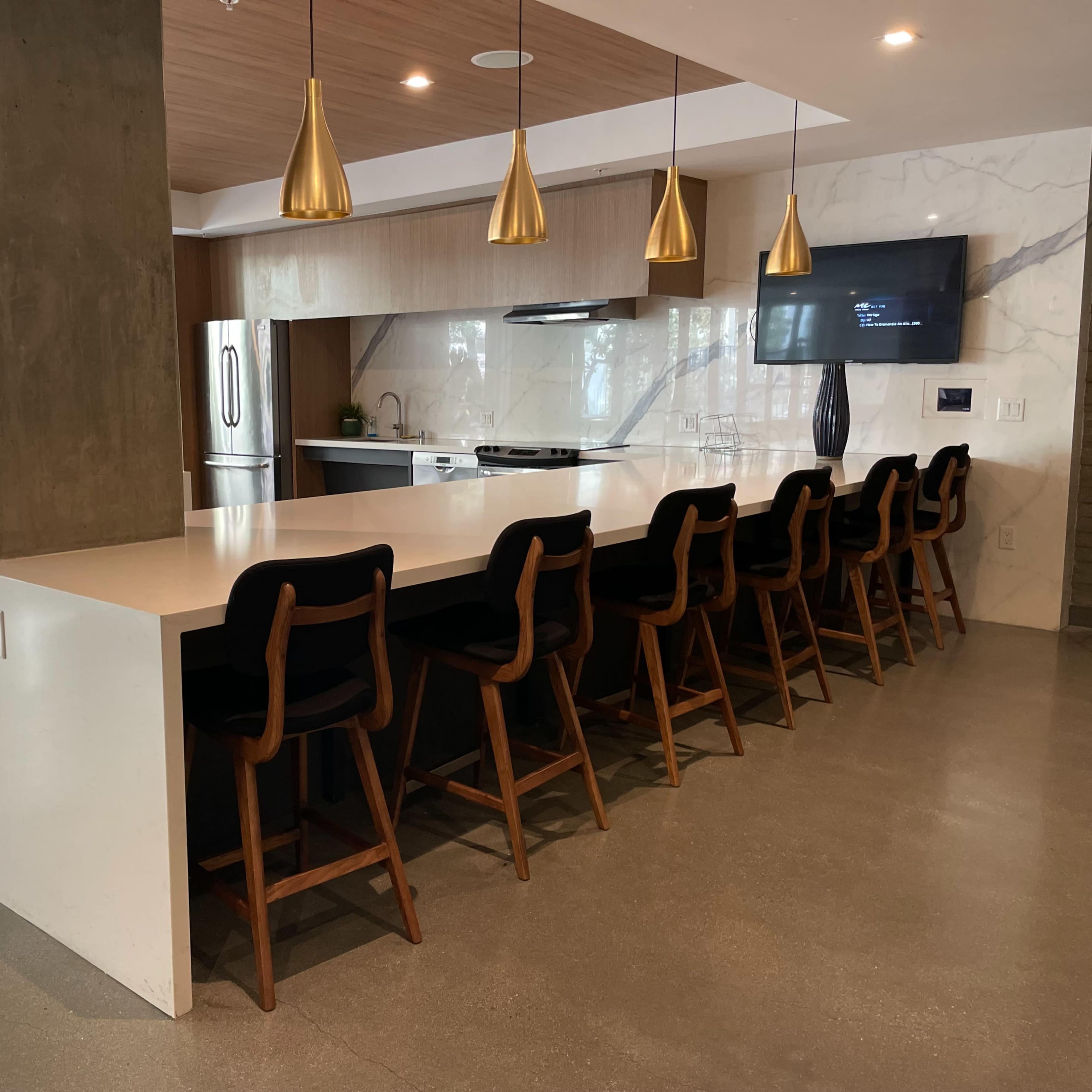 A modern kitchen with a long island bar and eight wooden stools, featuring pendant lighting and a large window overlooking a greenery-filled area.