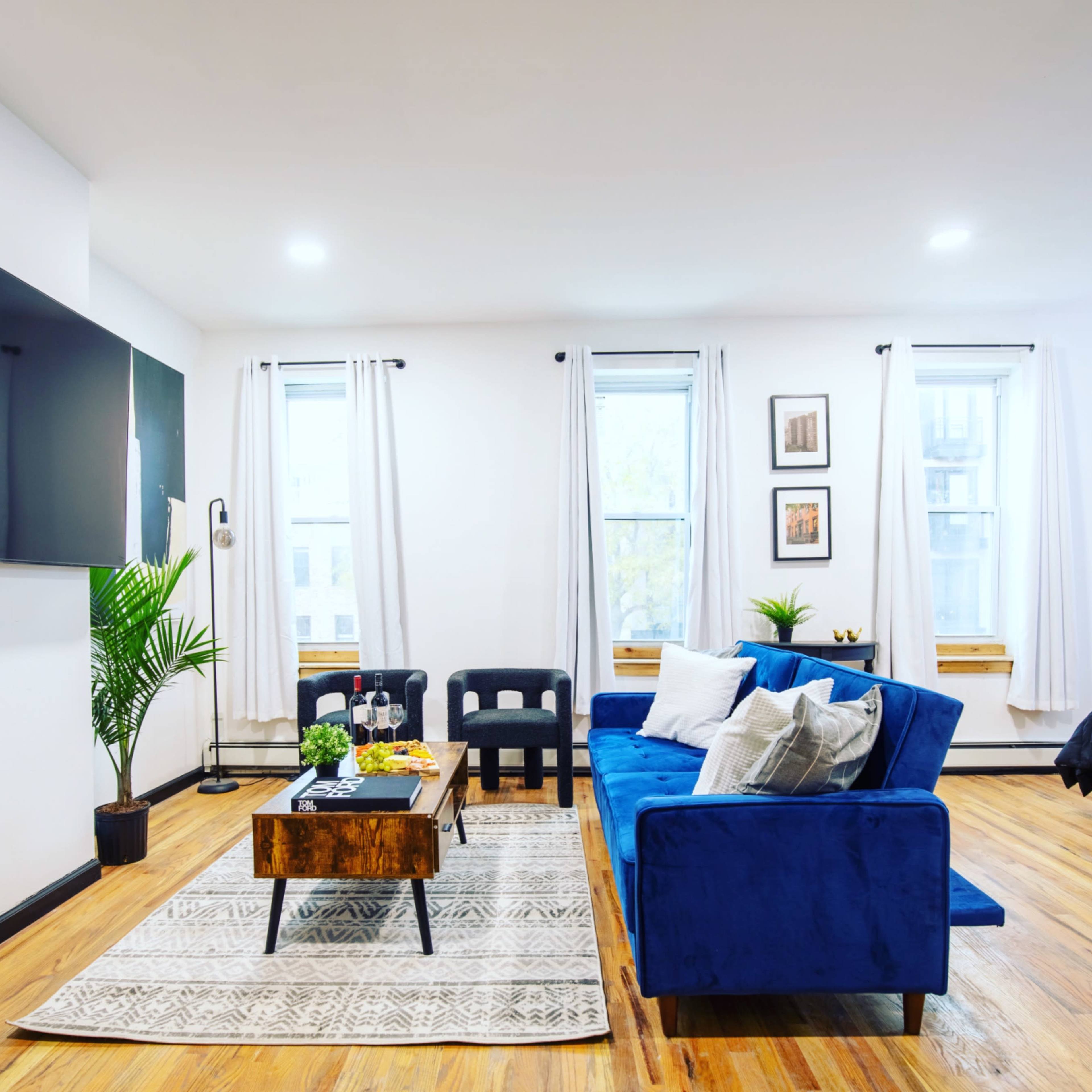 A modern living room features a blue sofa, a wooden coffee table, and a large television mounted on the wall, with windows allowing natural light to fill the space.