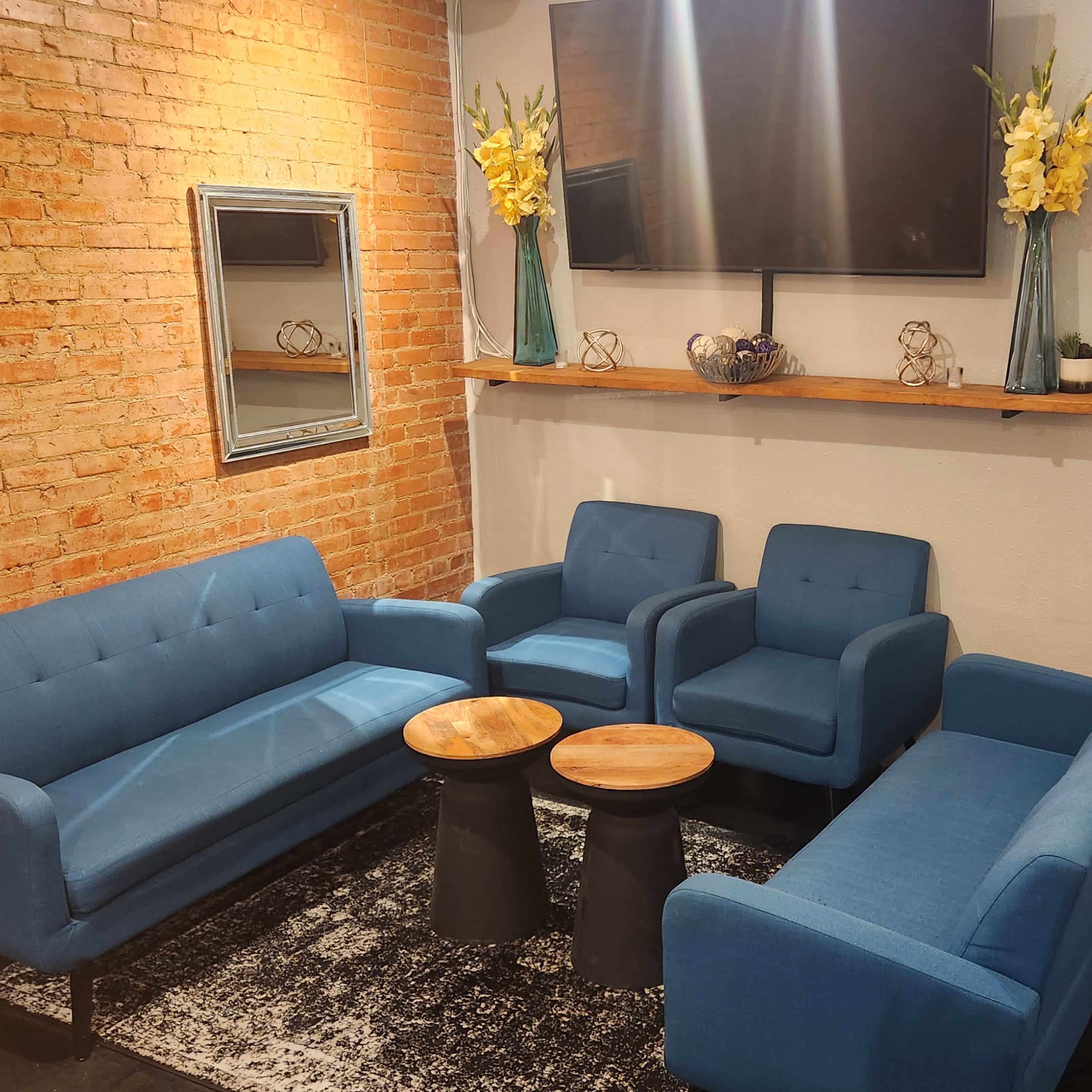 A cozy seating area with blue sofas and armchairs arranged around two small tables, against a backdrop of exposed brick walls and a mounted television.