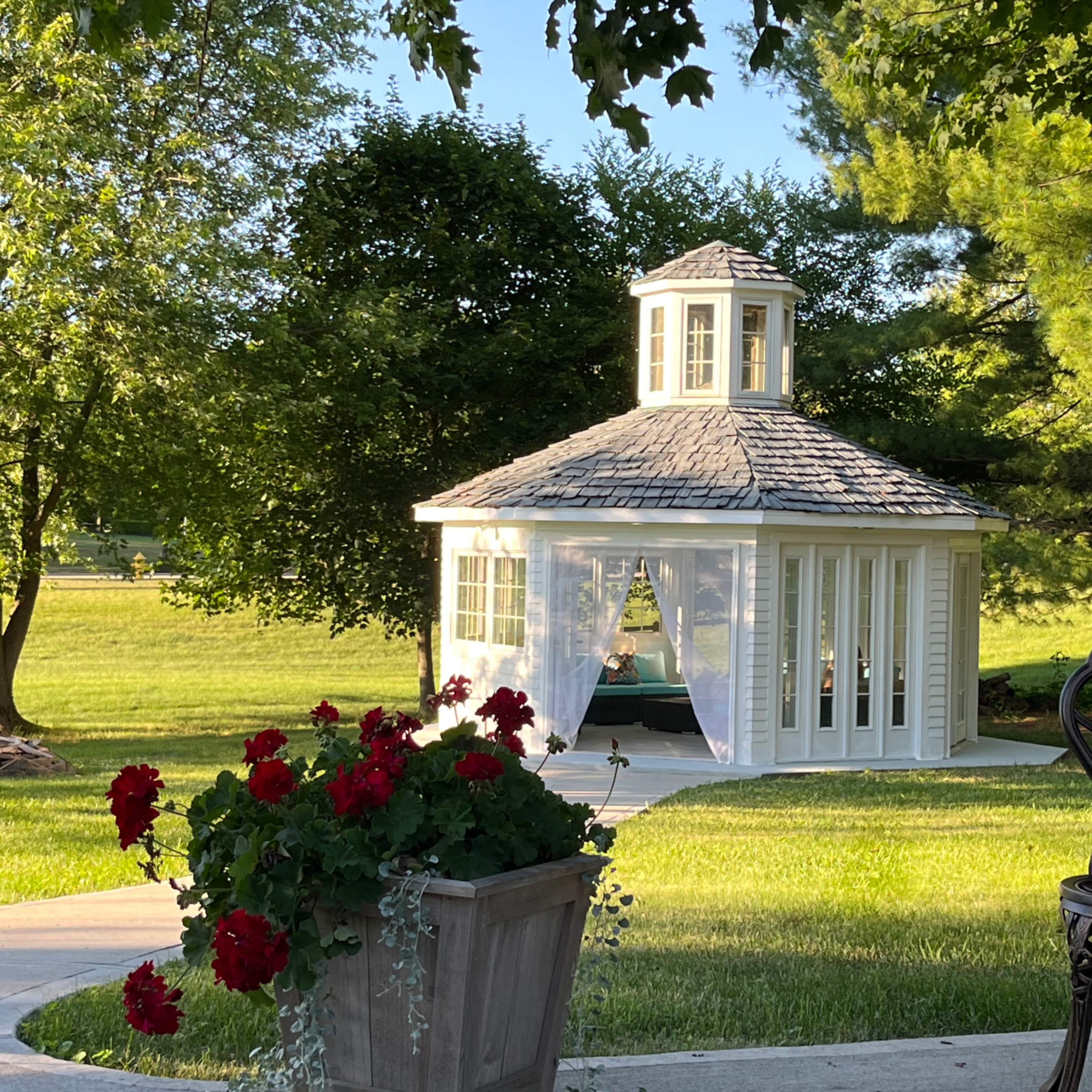 A white gazebo with a shingled roof is surrounded by green grass and trees, while red flowers bloom in a planter in the foreground.