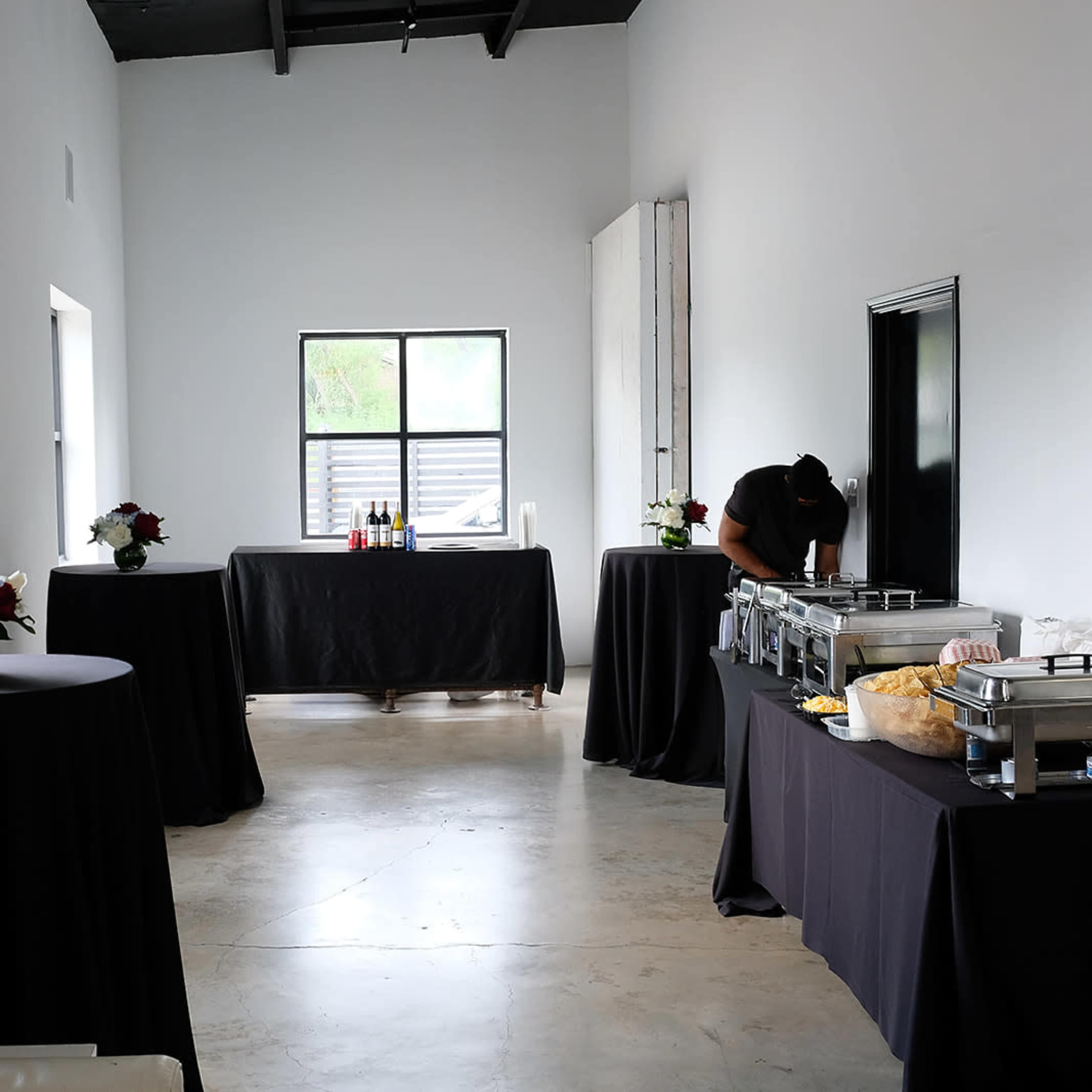A spacious room with two tables covered in black cloth, featuring food trays and floral arrangements, and a person preparing food at one end.
