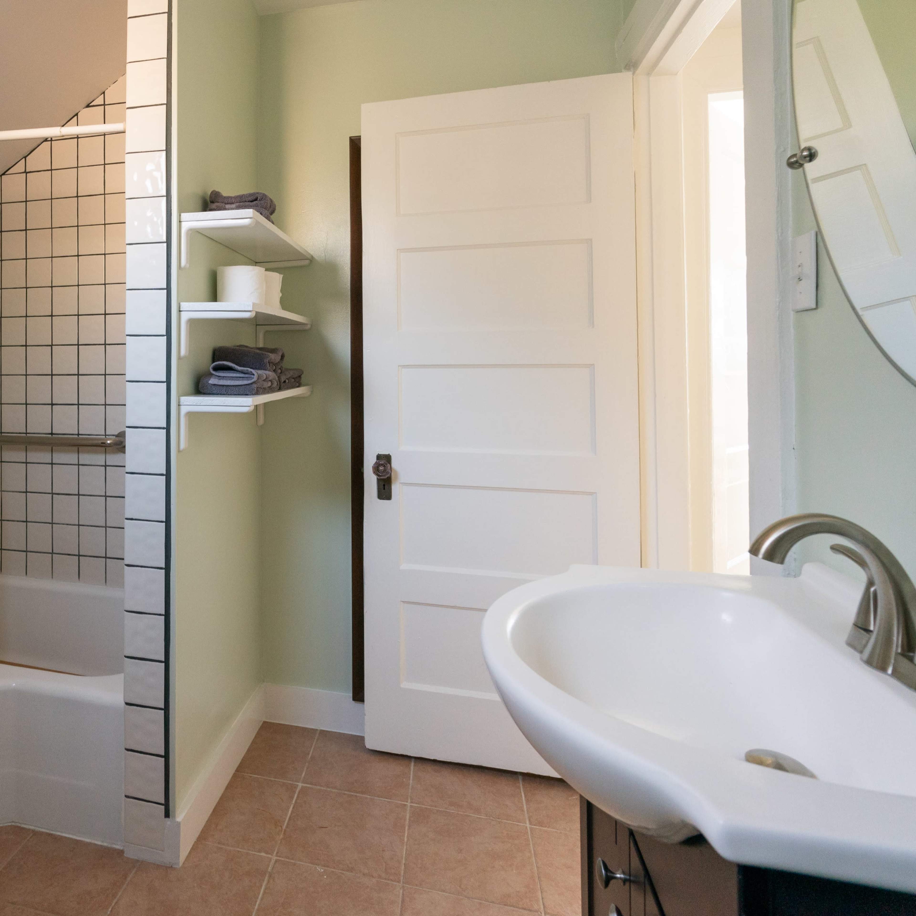 A bathroom featuring a shower/bath combination, shelves with towels, and a sink with a mirror against a light green wall.