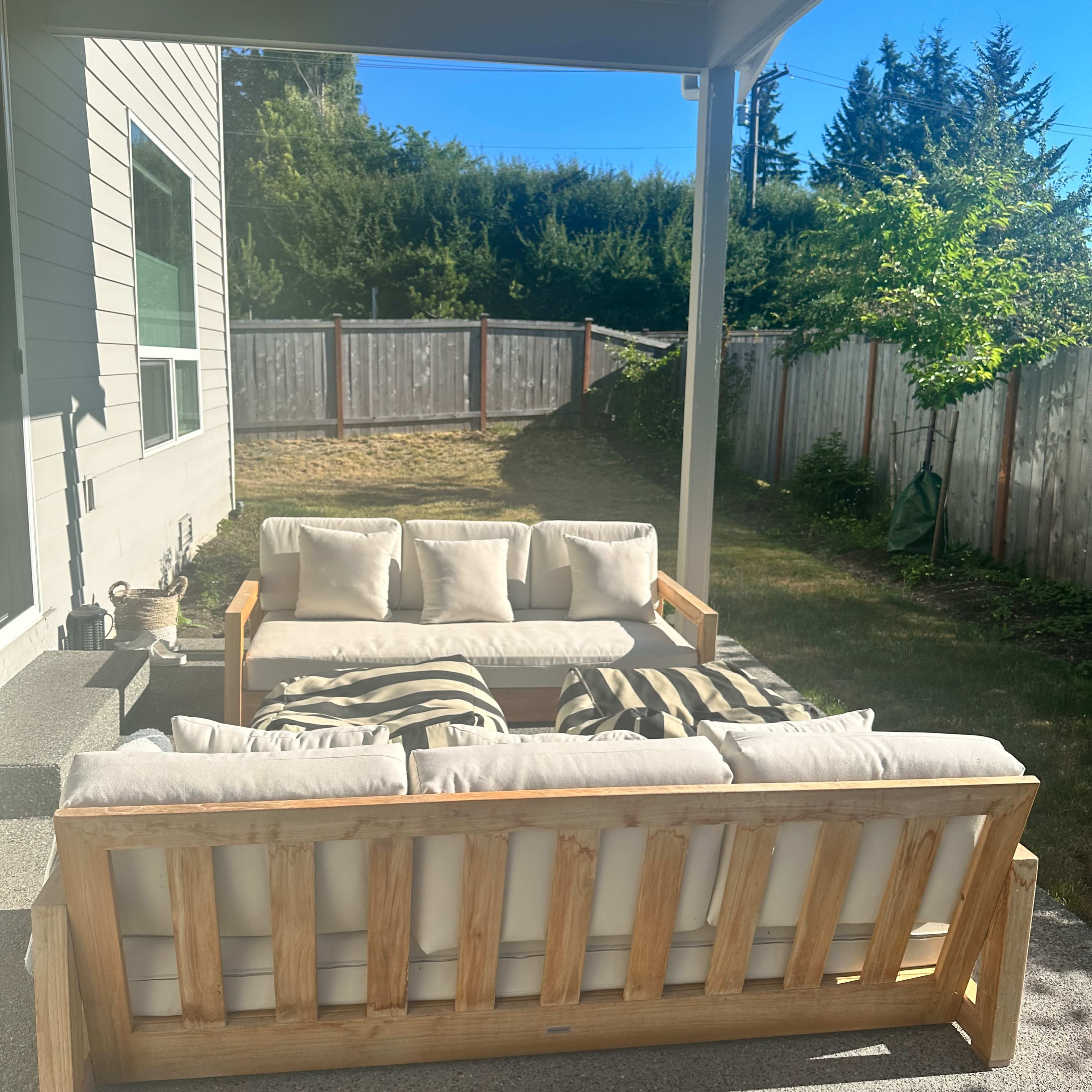 A wooden outdoor sofa with light-colored cushions is positioned under a covered porch, facing a grassy backyard surrounded by trees and a wooden fence.