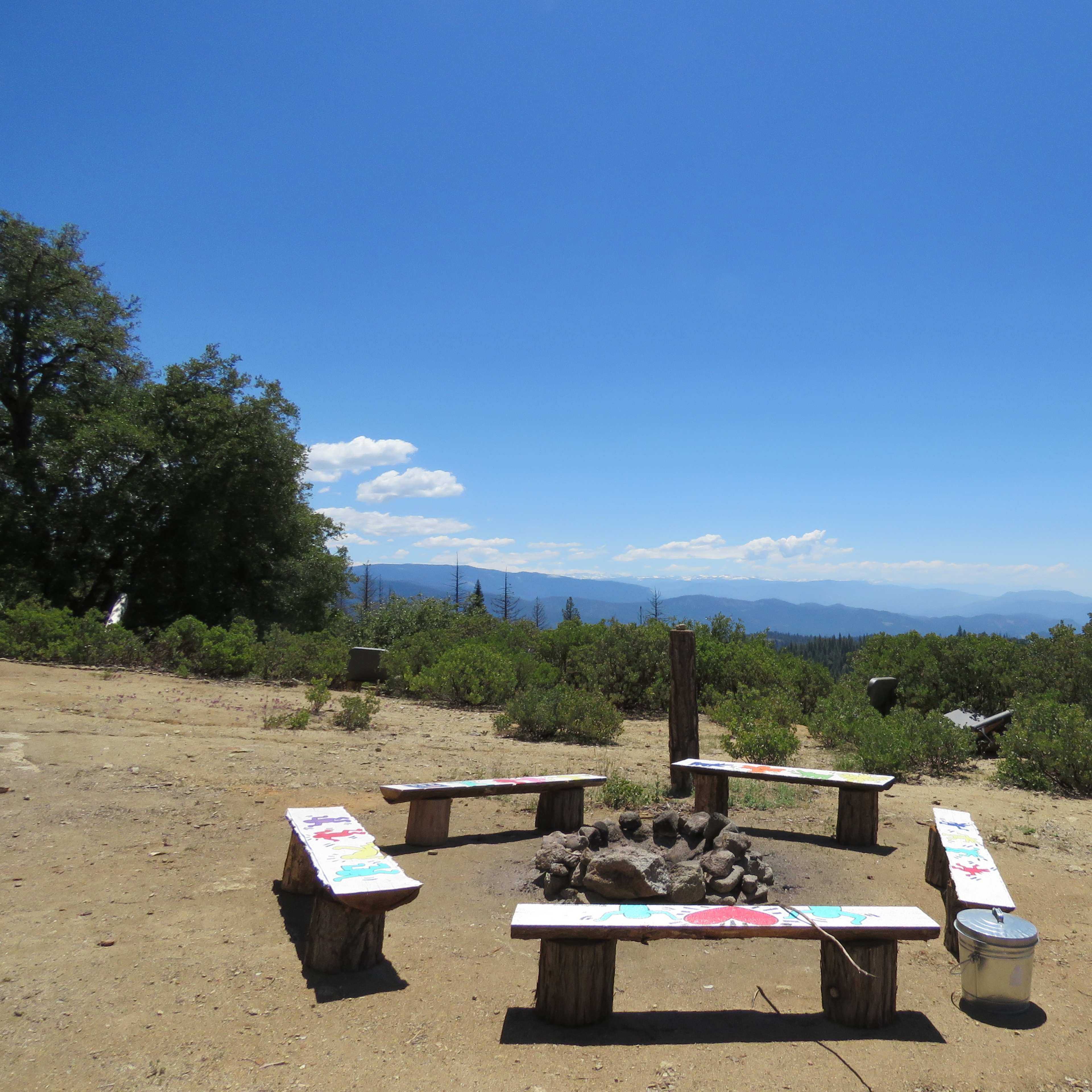 A campfire circle with benches around a fire pit, set against a clear blue sky and mountain landscape.