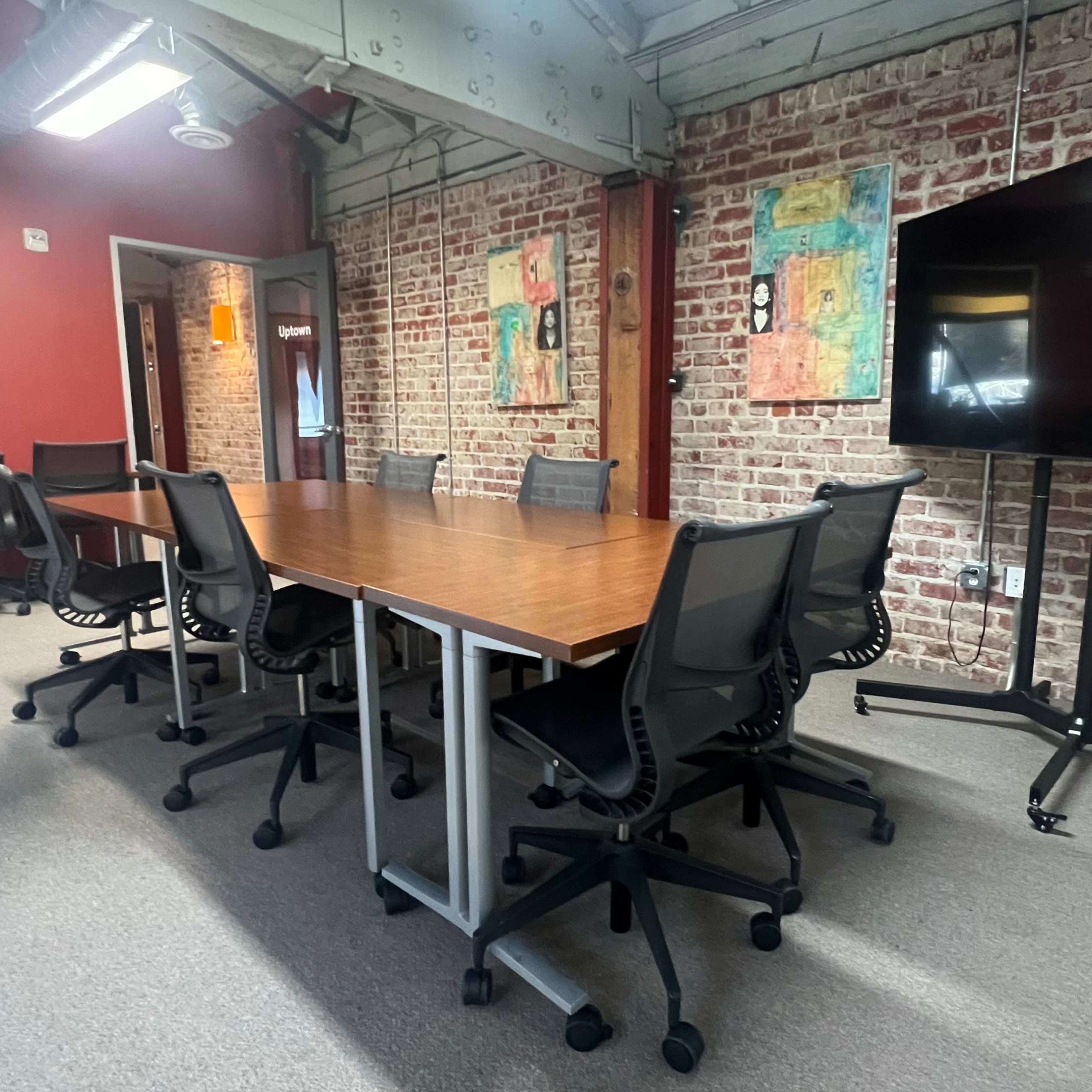 A conference room features a large wooden table surrounded by black office chairs, with exposed brick walls and a large screen on a stand.