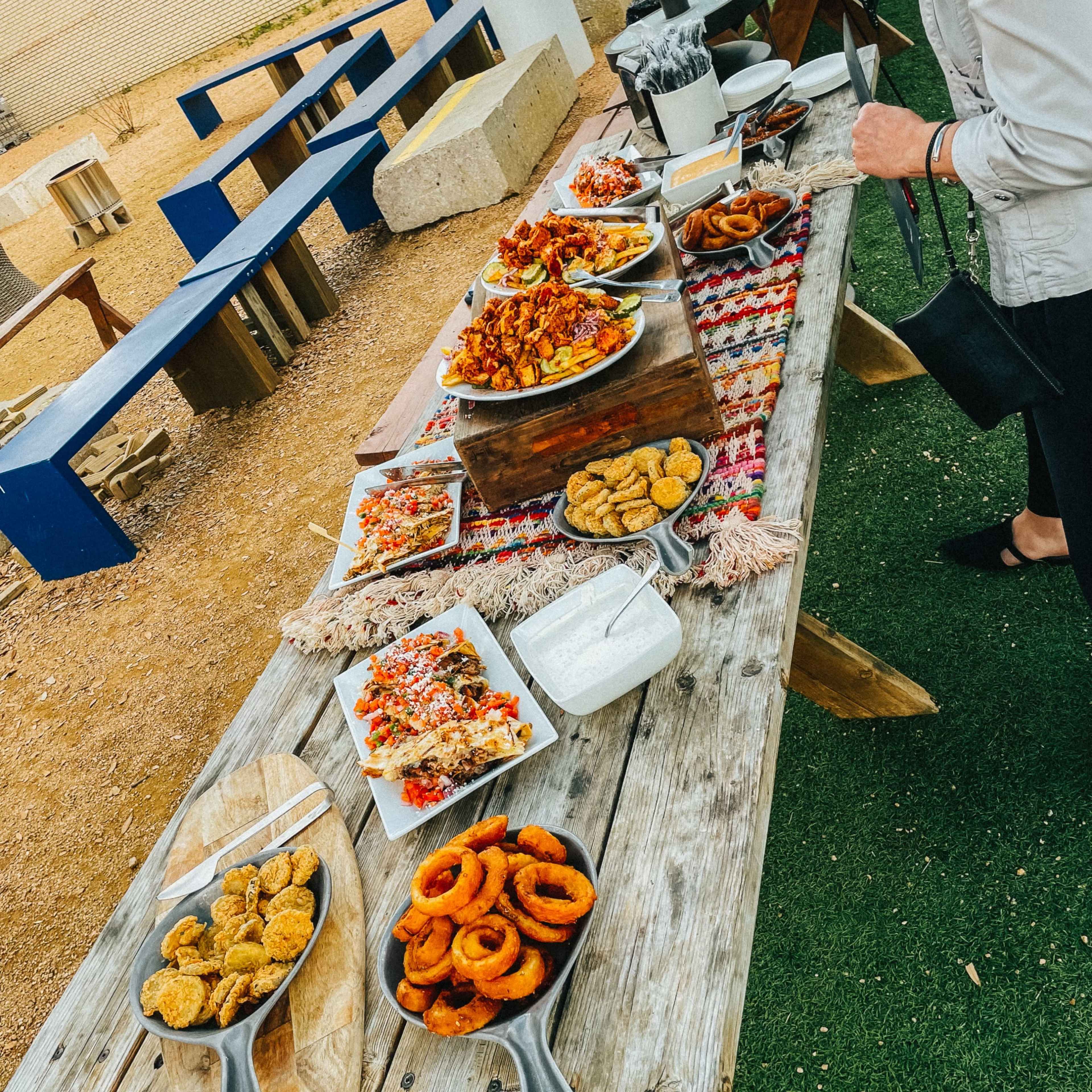A wooden table is filled with various platters of food, including fried items, salads, and sauces, set in an outdoor area with benches nearby.
