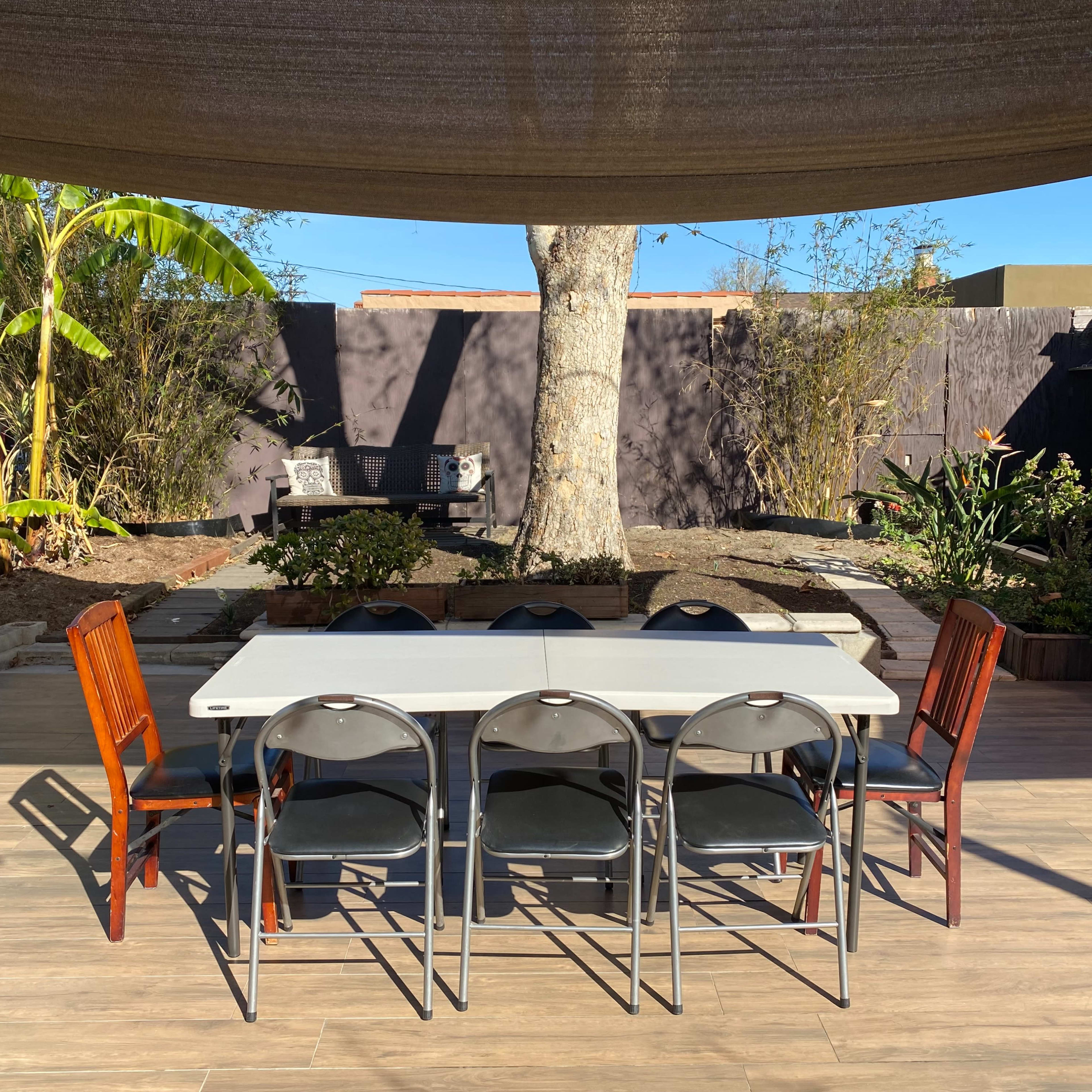 A rectangular table surrounded by six chairs is set in a backyard with a large tree and landscaped greenery in the background.