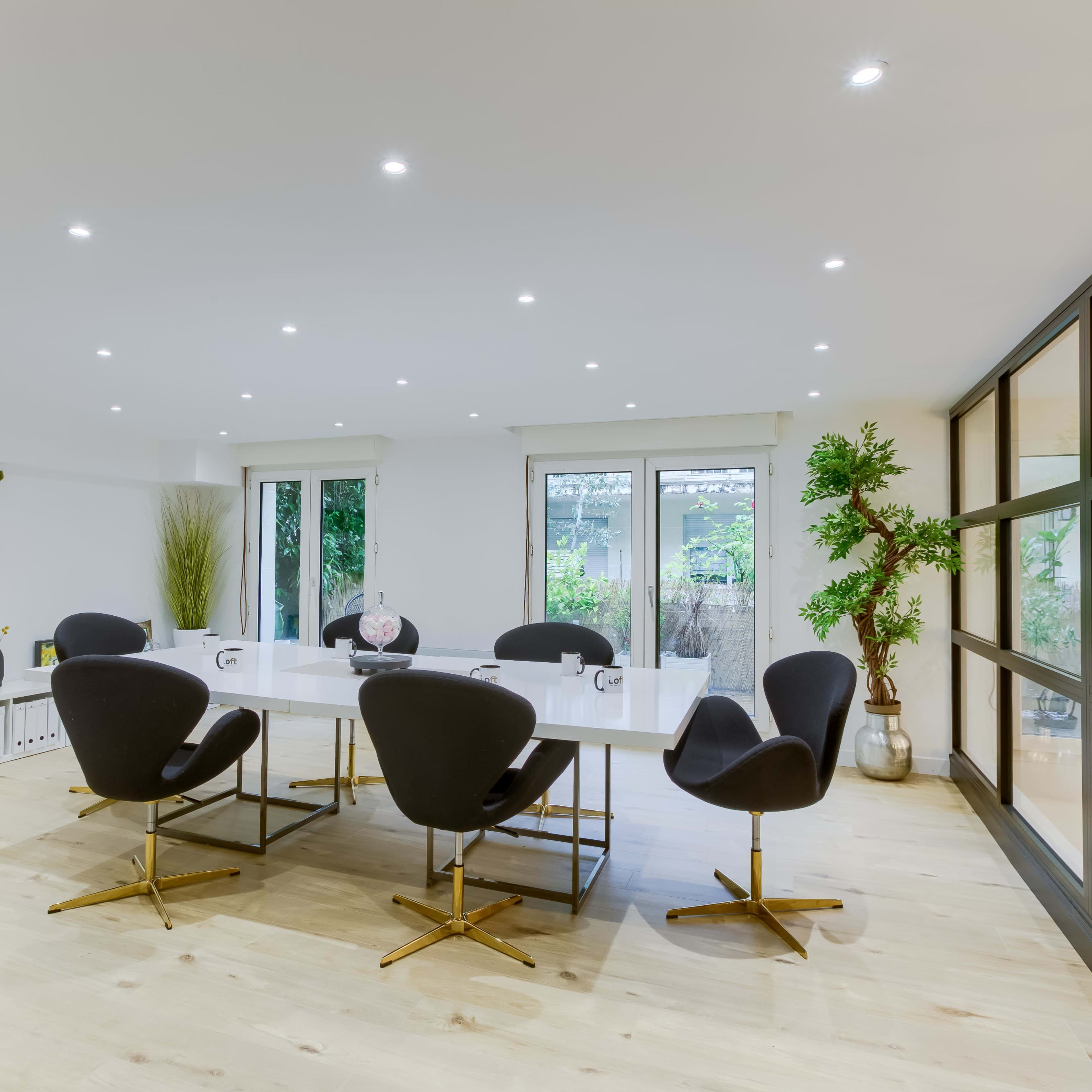 A modern conference room features a large white table surrounded by black chairs, with glass doors leading to an outdoor area and greenery in the decor.