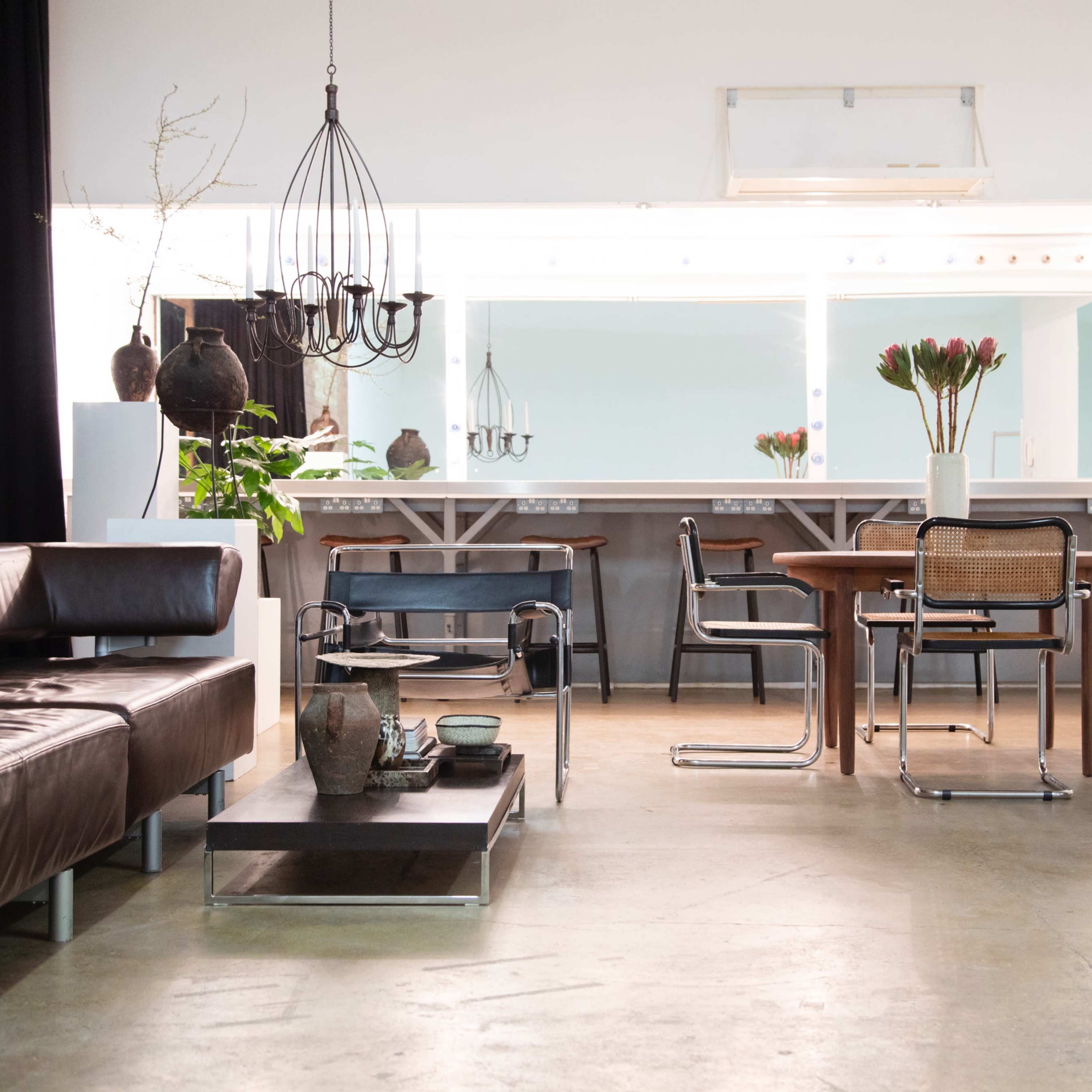 A modern interior space featuring a leather sofa, a dining table with chairs, and a kitchen area with a large mirror and decorative plants.