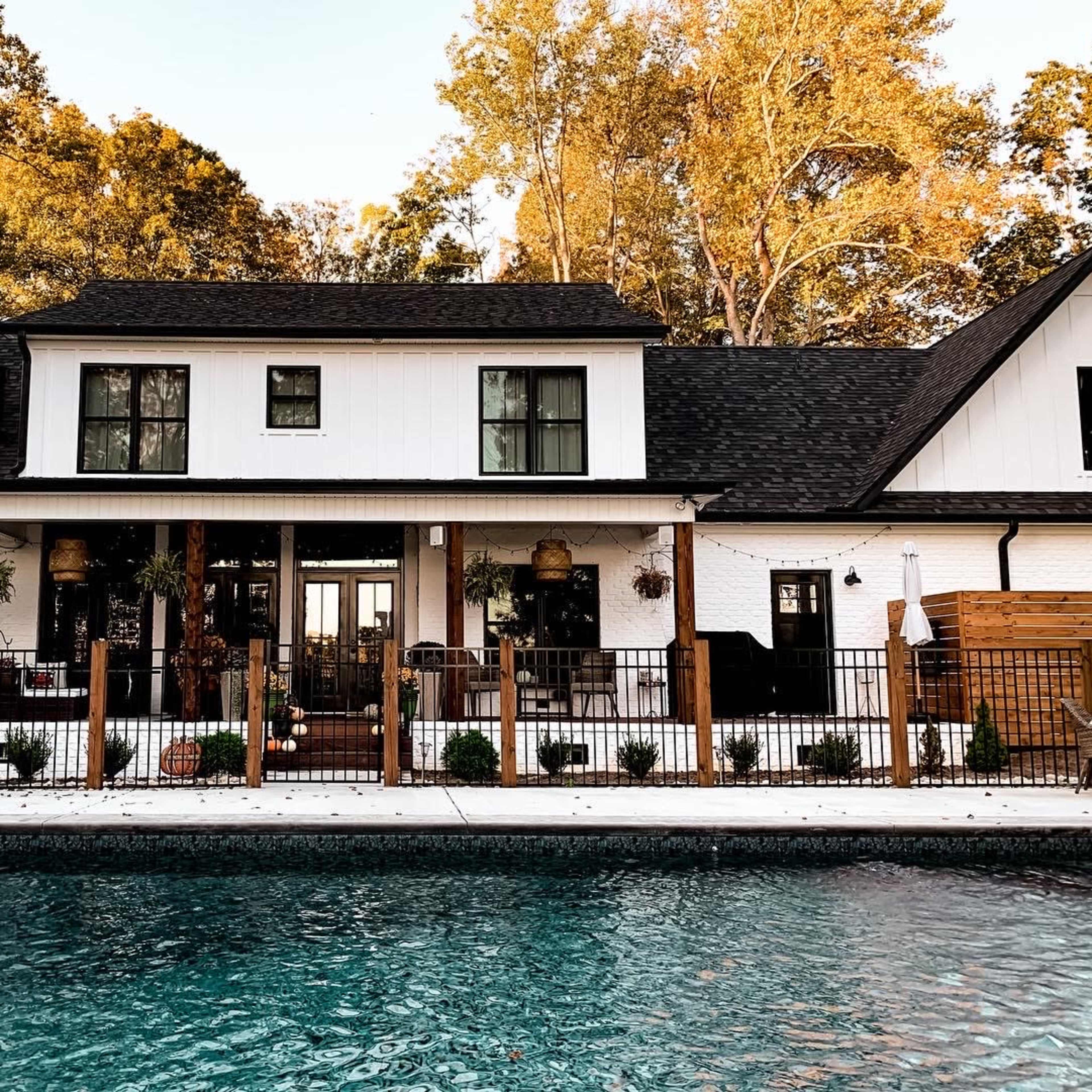 A two-story, modern farmhouse with a black roof and white exterior features a fenced patio area in front of a swimming pool.