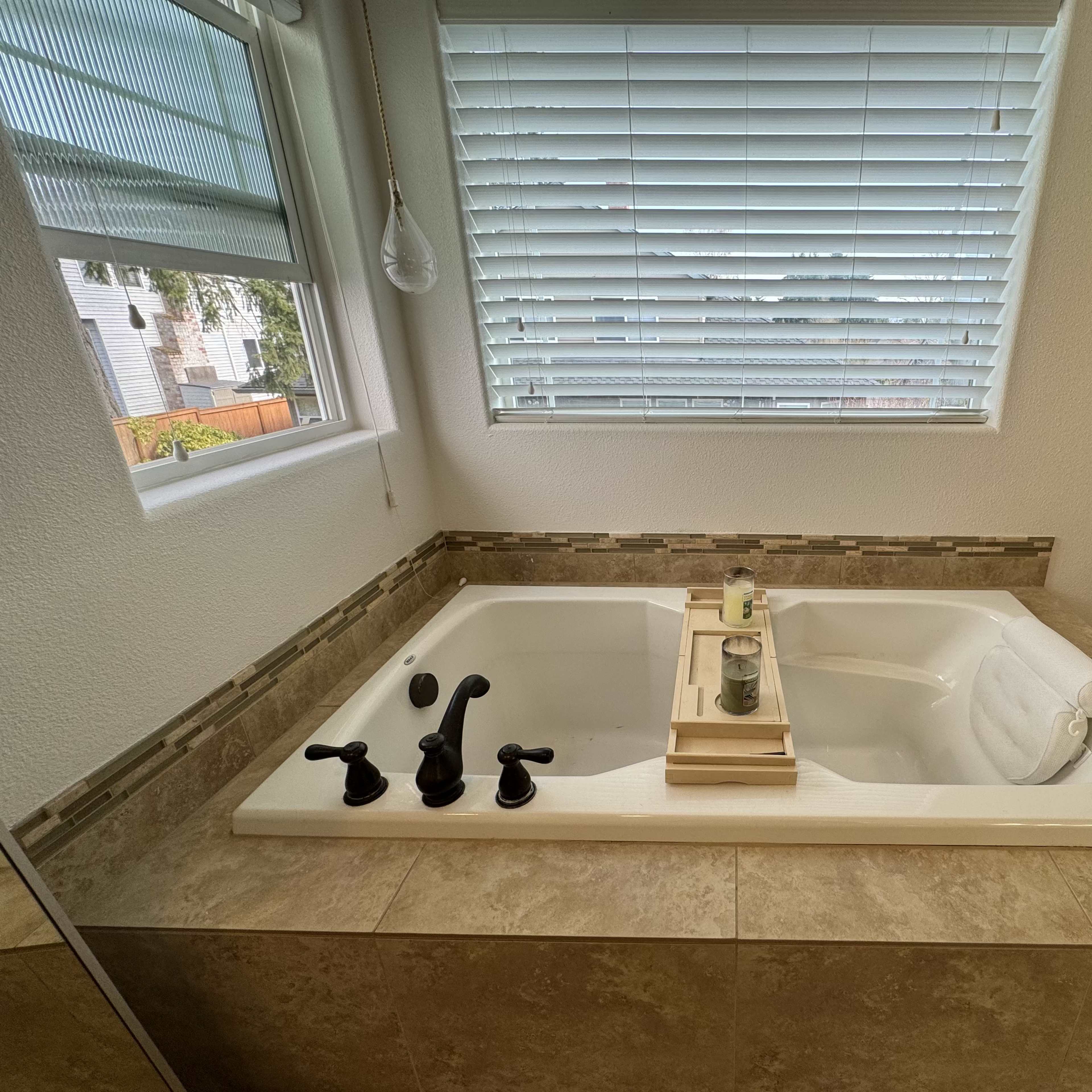 A modern bathroom featuring a jetted bathtub with a wooden tray holding candles, set against a window with blinds.