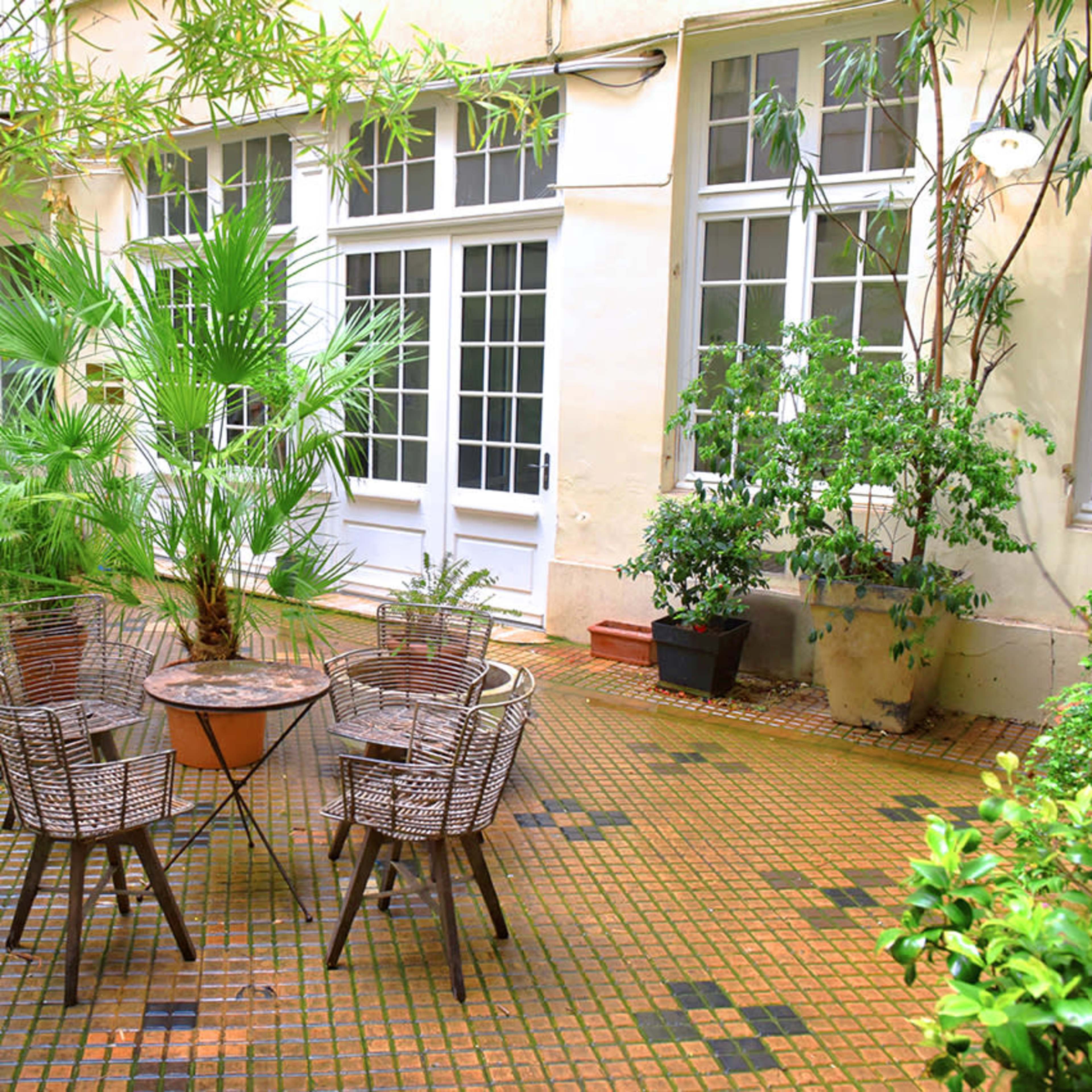 A small courtyard features a circular table surrounded by four woven chairs, set among various potted plants and greenery.