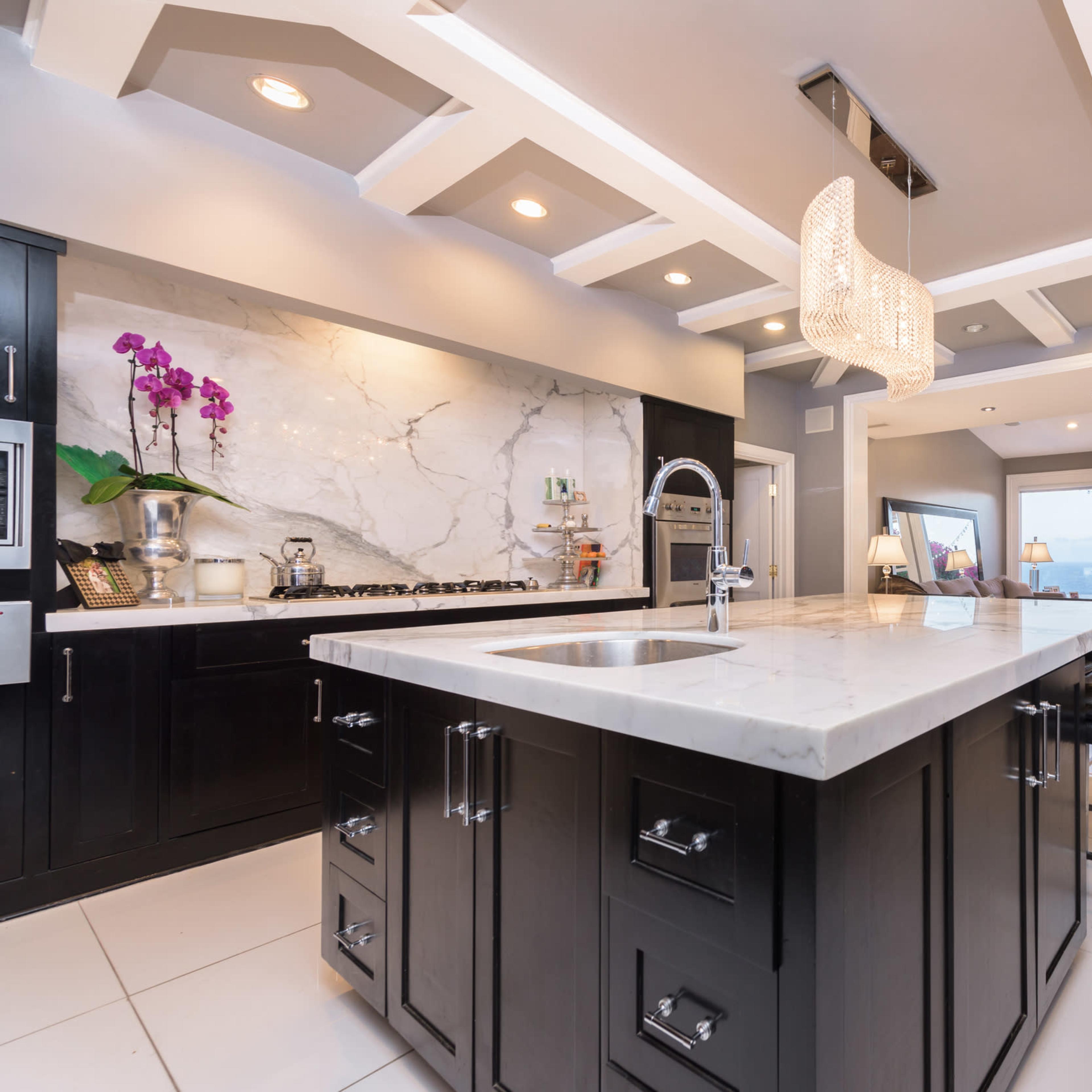 A modern kitchen featuring a large marble island, dark cabinetry, and an elegant chandelier above.