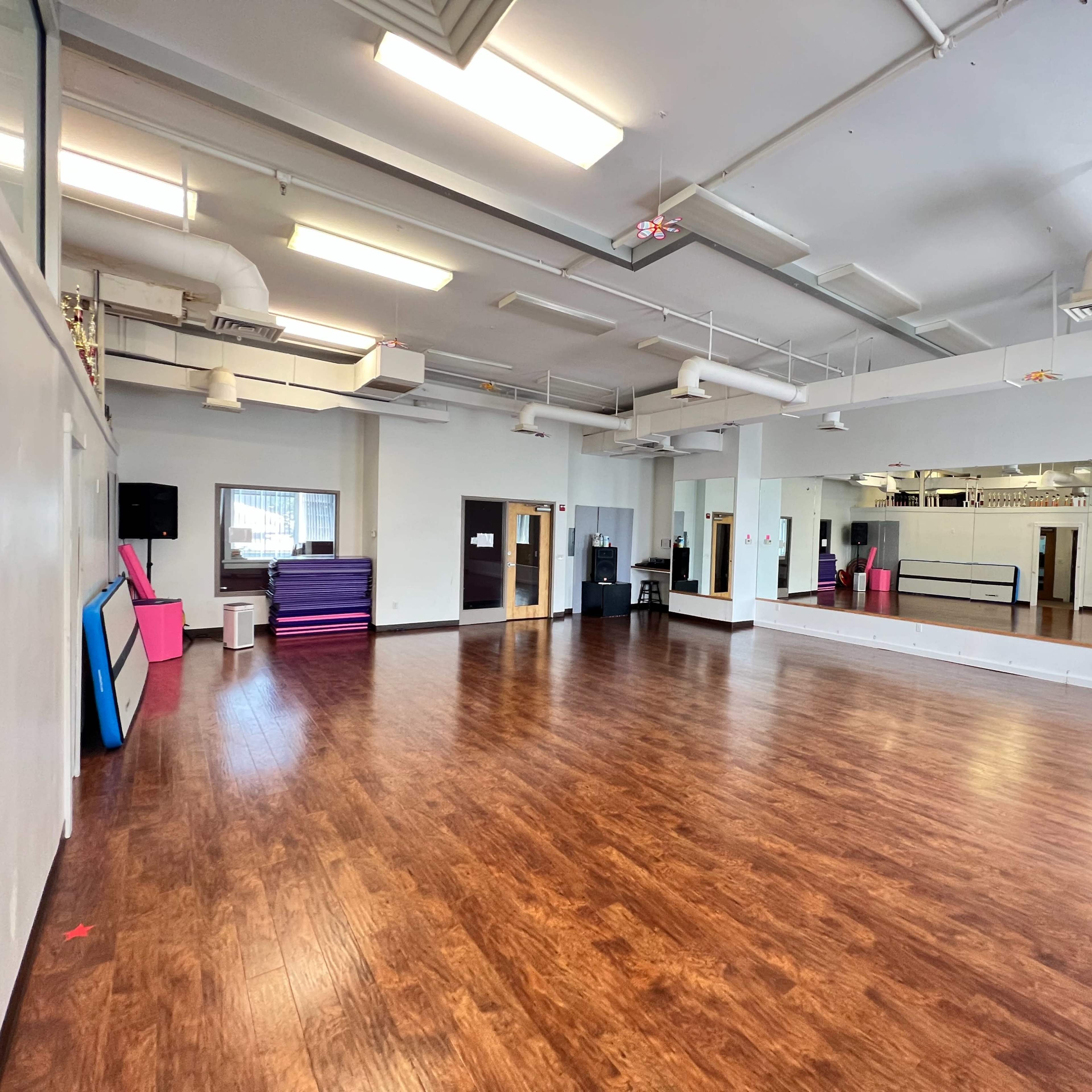 A spacious dance studio with hardwood flooring, mirrors on one wall, and colorful mats stacked in the corner.
