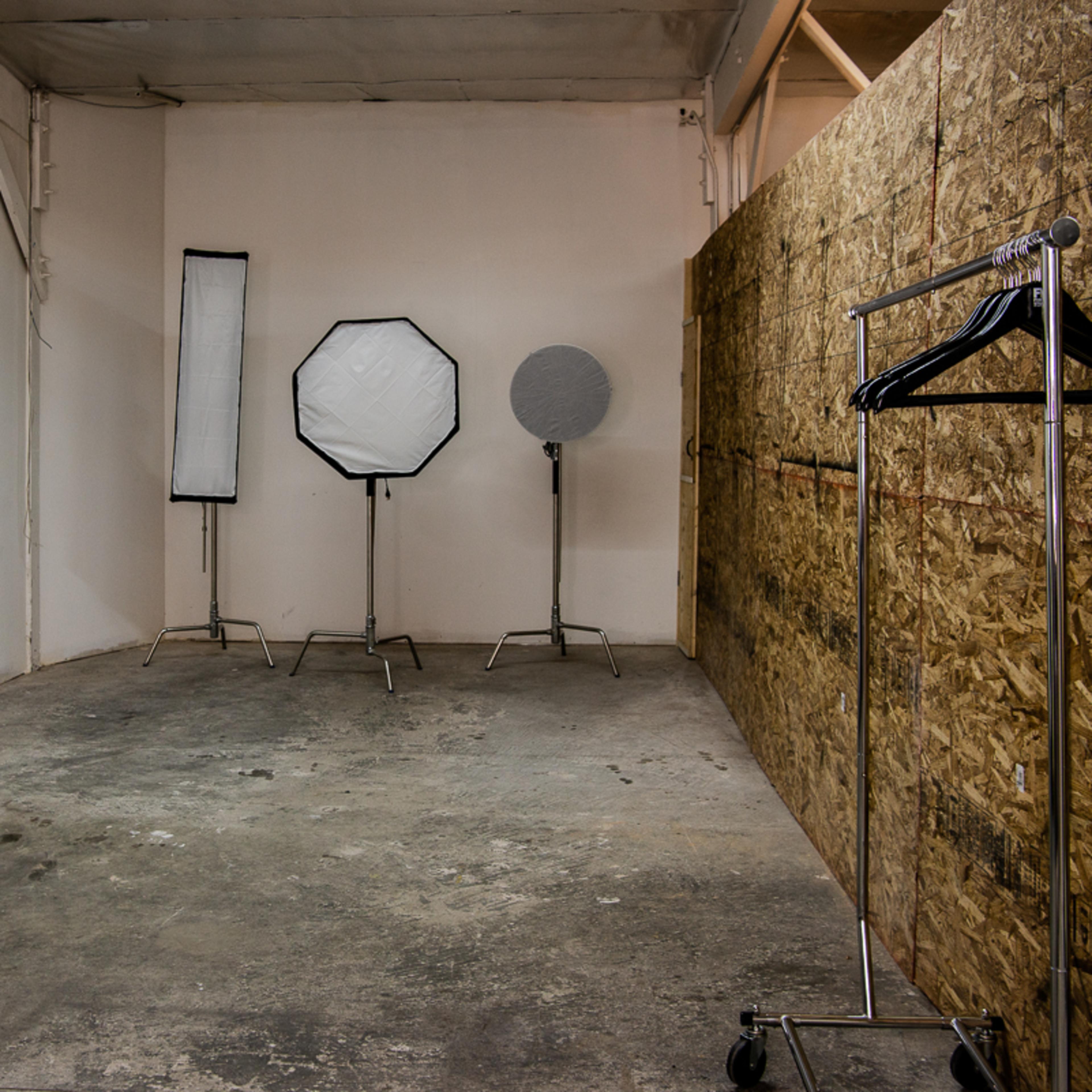 An empty photography studio with three lighting equipment stands and a clothing rack against a textured wall.
