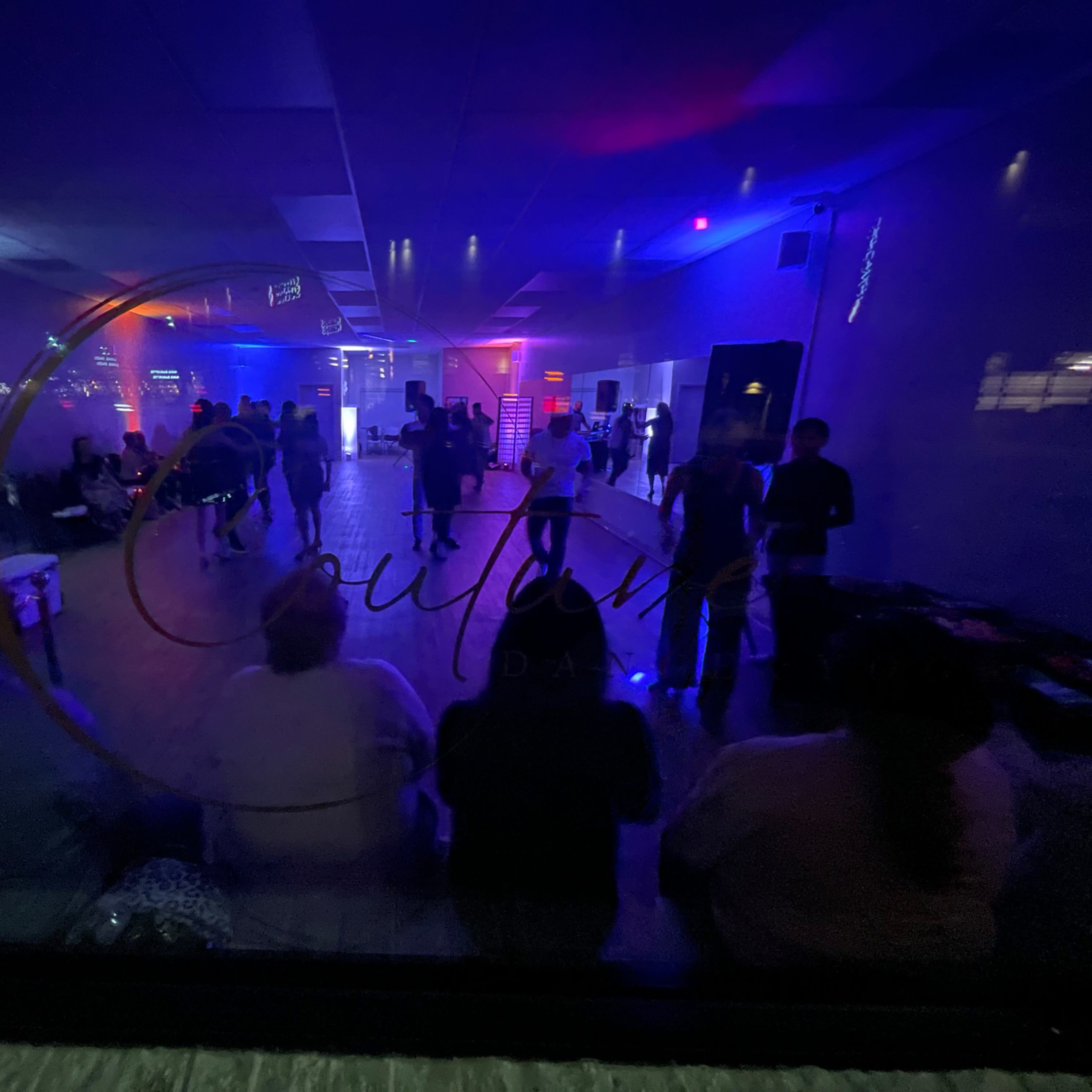 A dimly lit dance hall with people interacting and colorful lights illuminating the space.