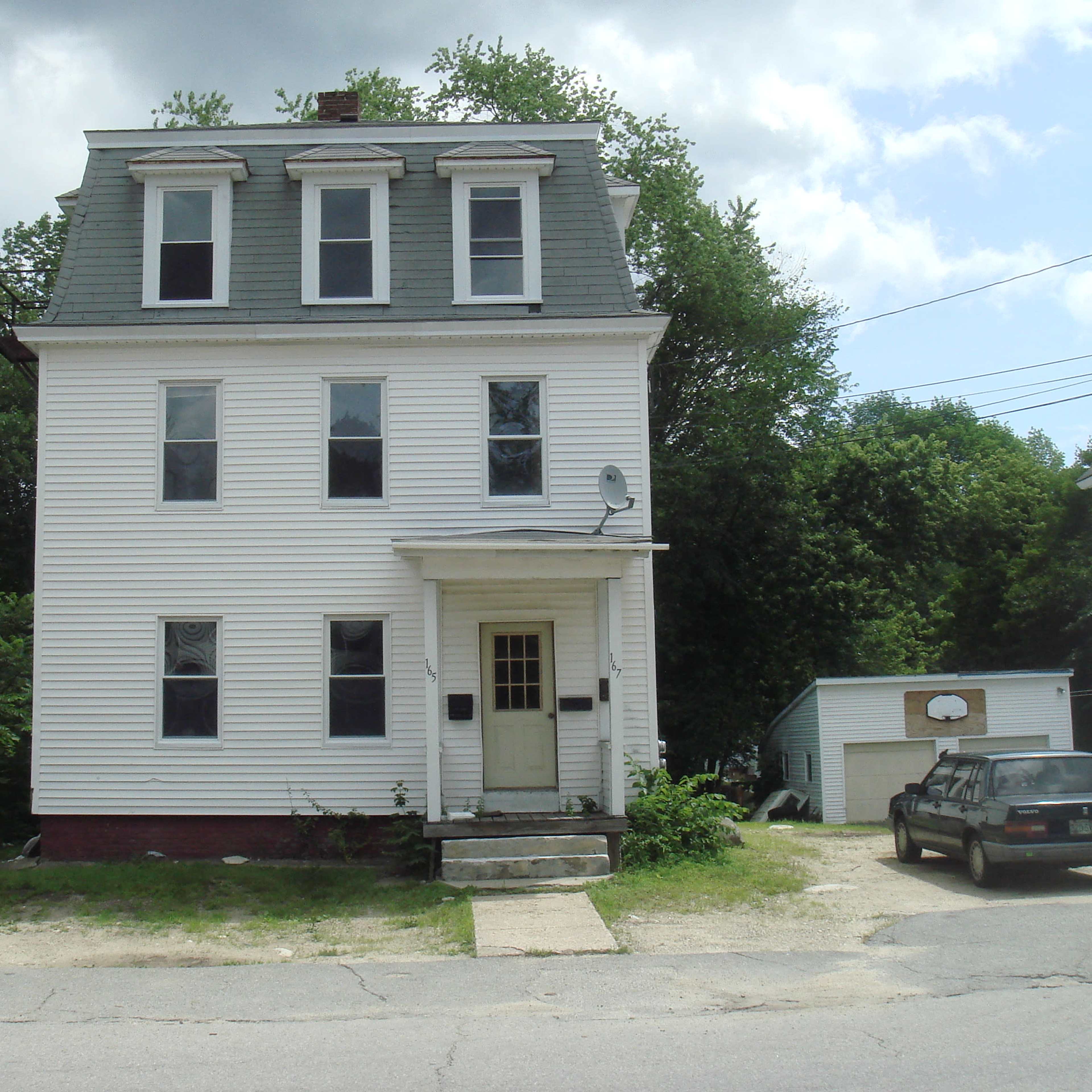A three-story white house with a sloped roof, located on a residential street, surrounded by greenery.