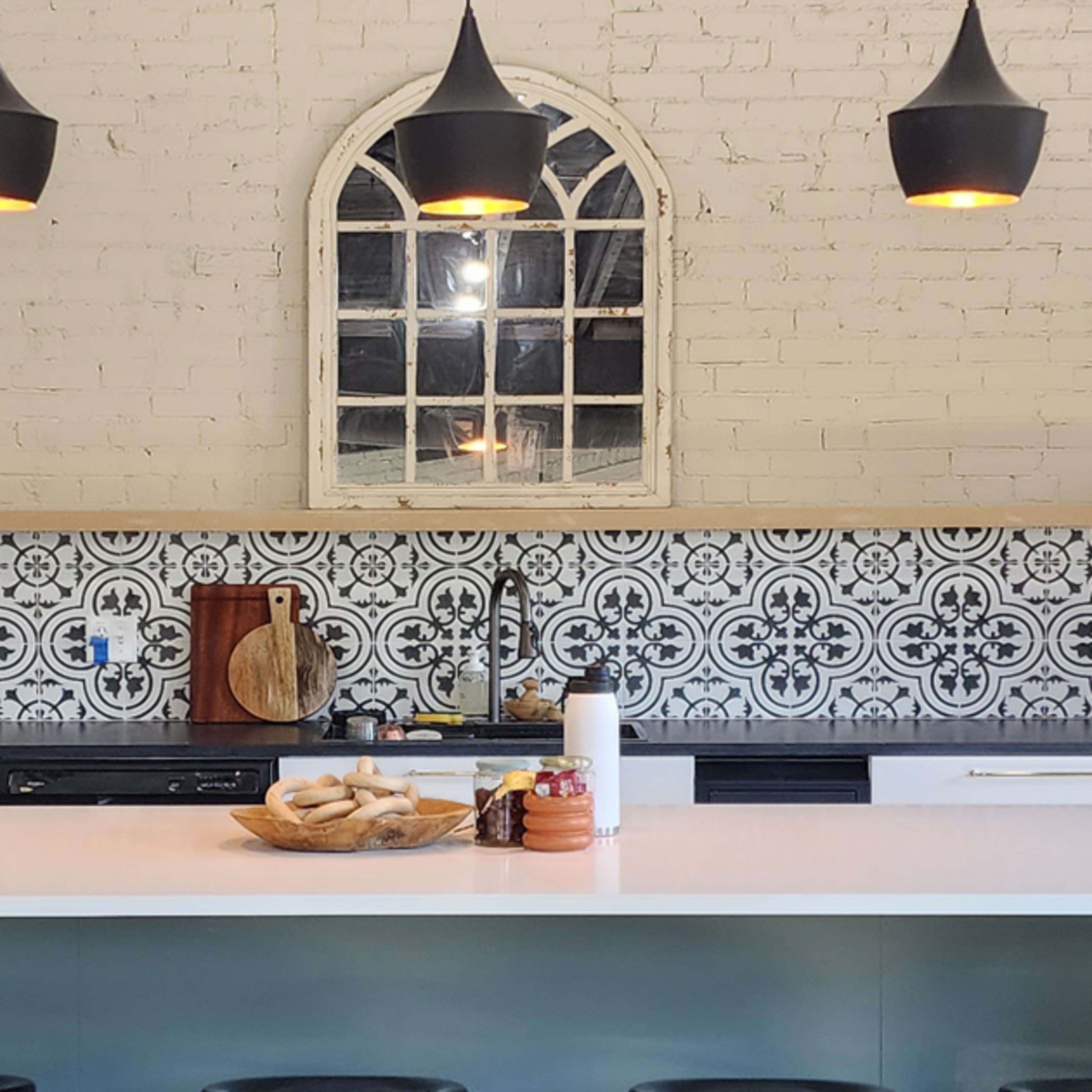 A modern kitchen features a white island with a wooden bowl of bread, black pendant lights, and a patterned backsplash.