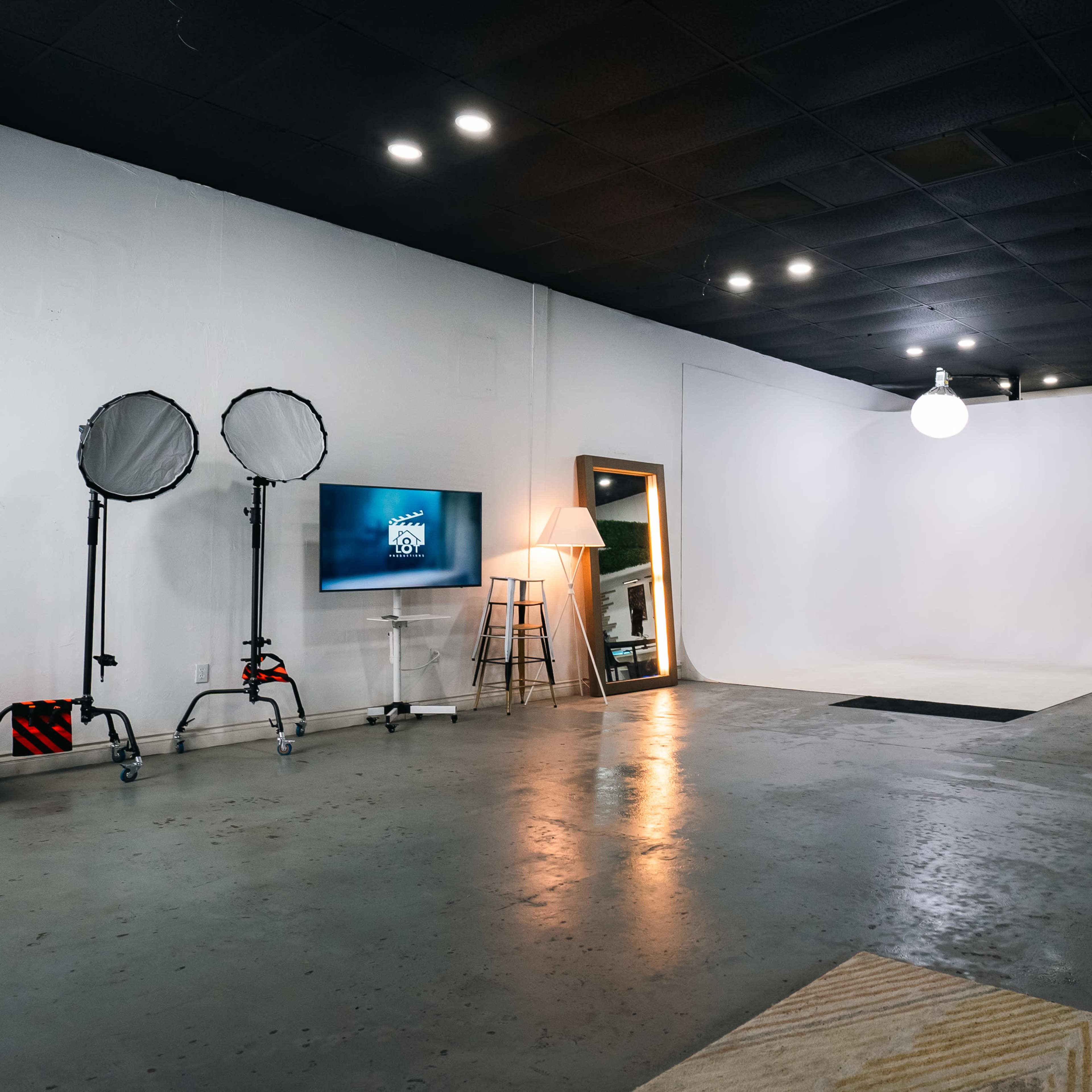 A spacious studio with a blank white backdrop, equipped with lighting stands, a television on a stand, and various furniture pieces.