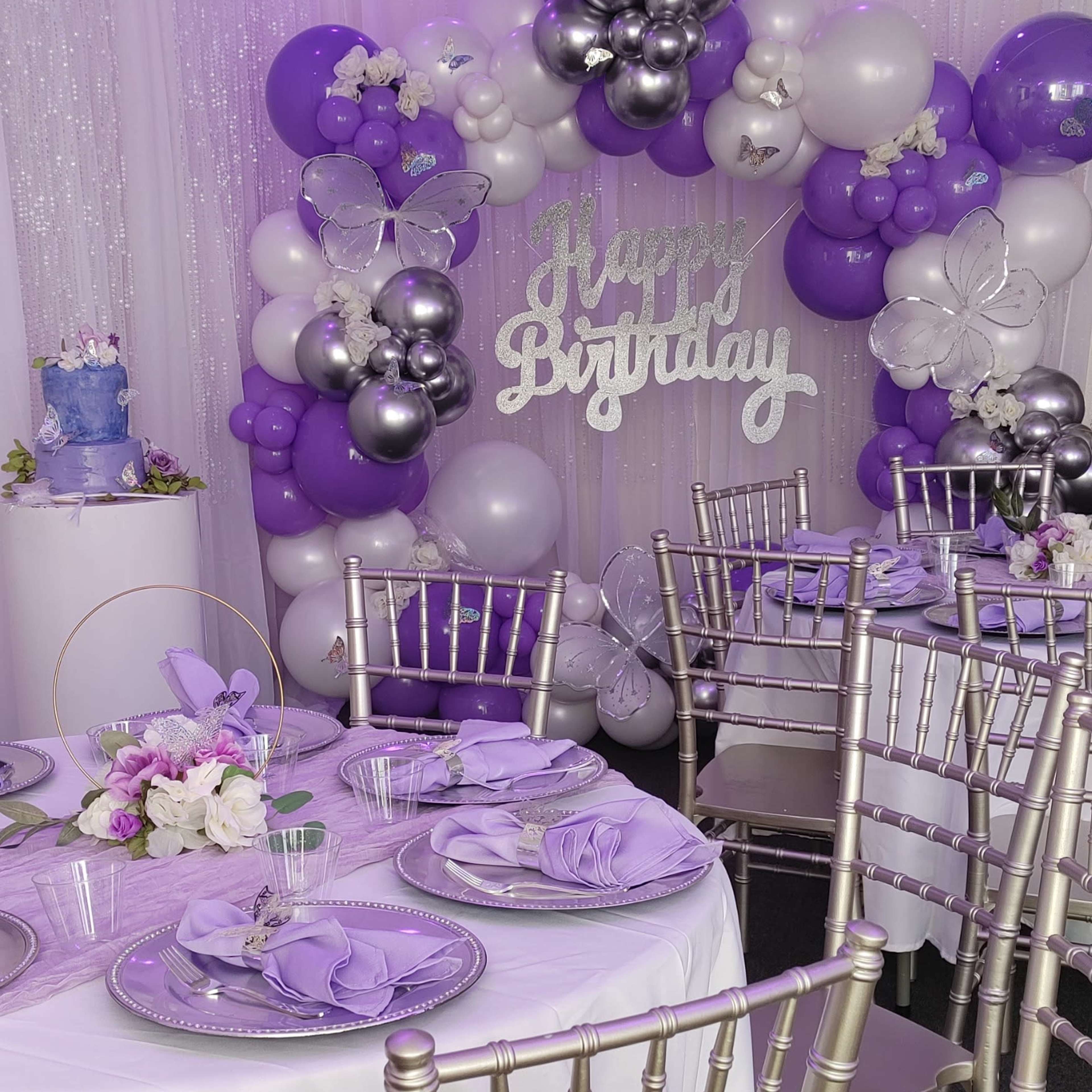 A birthday party setup with a table adorned in purple and silver decorations, featuring a cake and a backdrop of balloons.