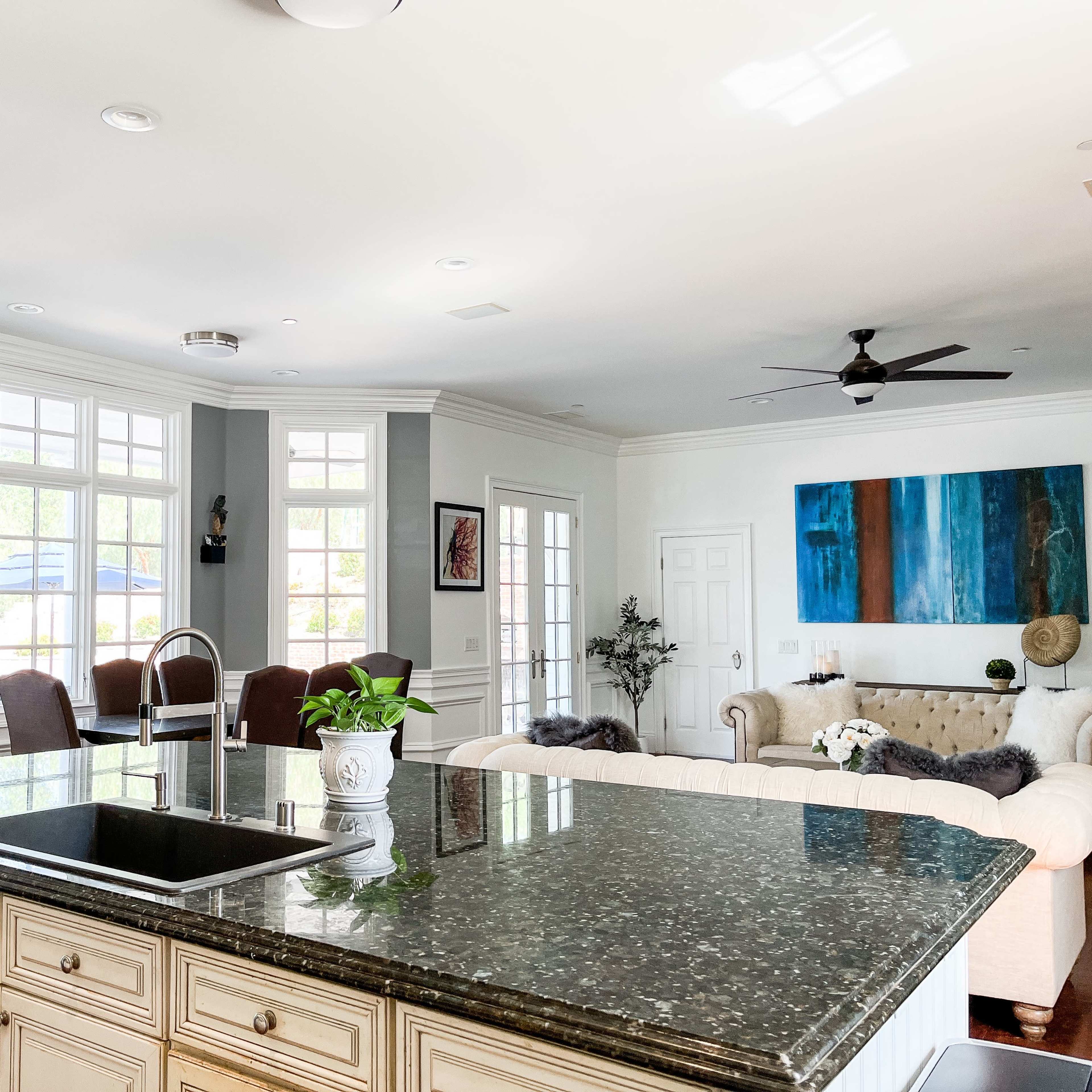 A modern kitchen with a dark granite island, light-colored cabinetry, and large windows providing natural light, overlooking a living area with a sofa and artwork on the walls.
