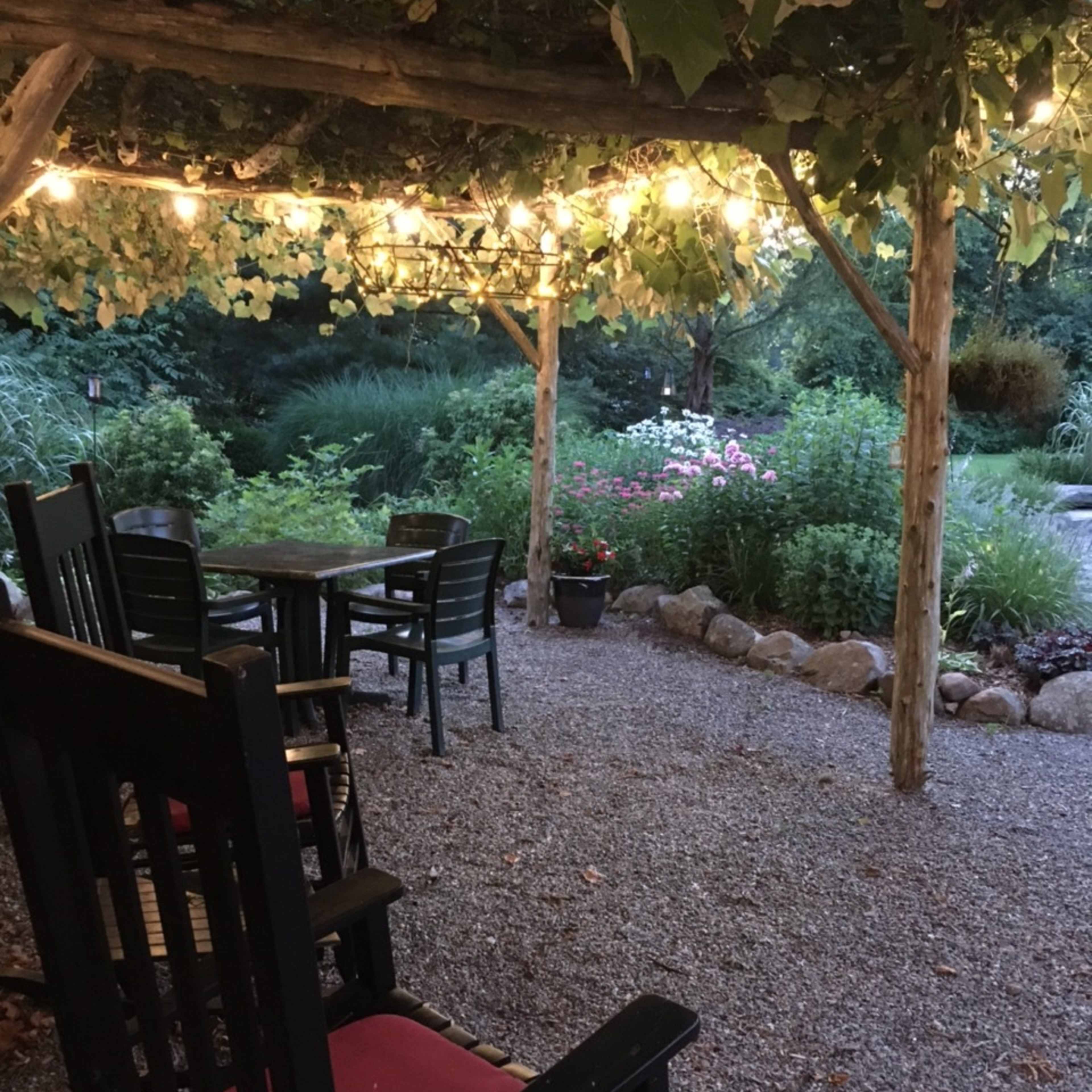 A gravel garden space features wooden rocking chairs and a table beneath string lights and a leafy arbor.