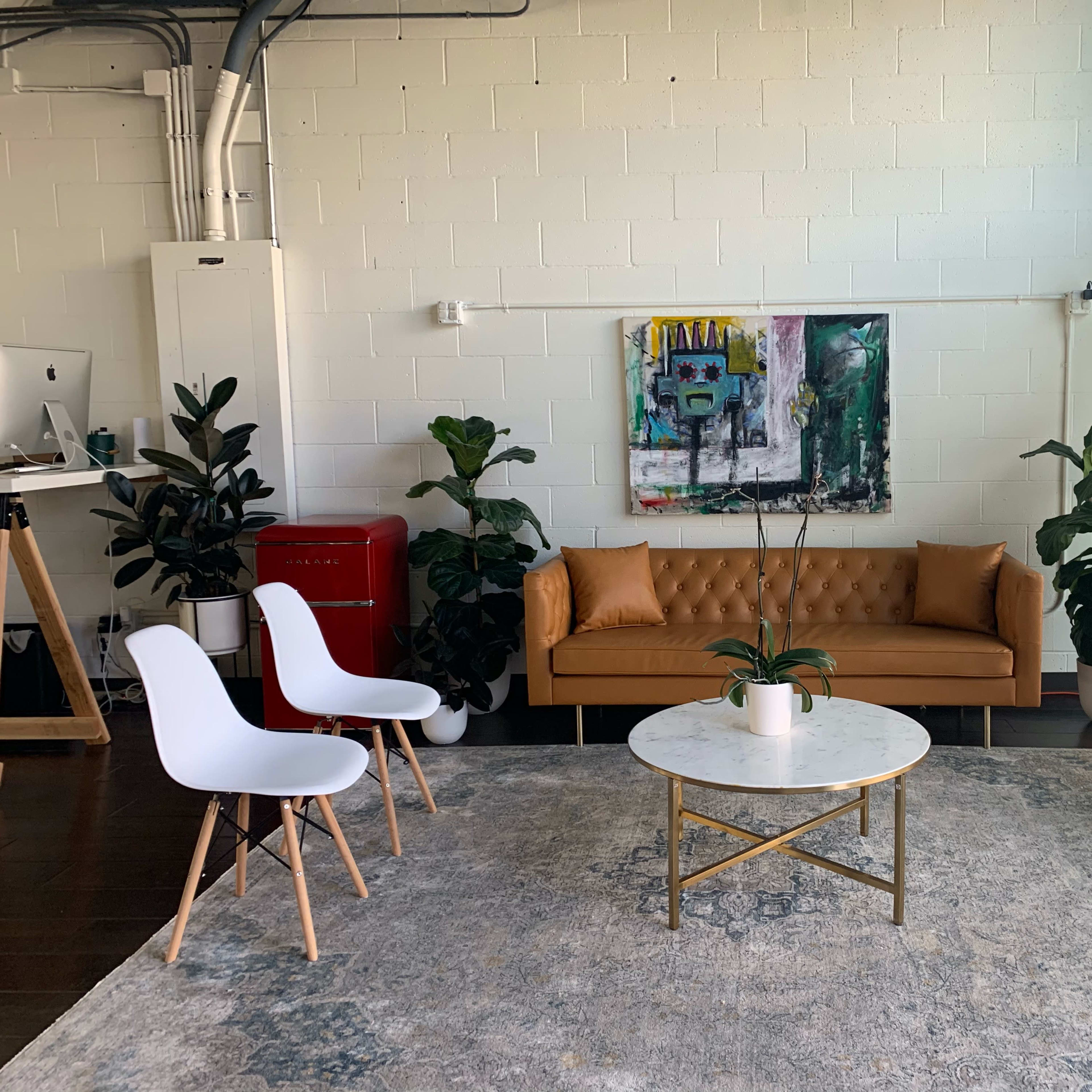A modern living space featuring a brown sofa, a round marble coffee table, two white chairs, and various plants, all set against a light-colored wall with a colorful artwork.