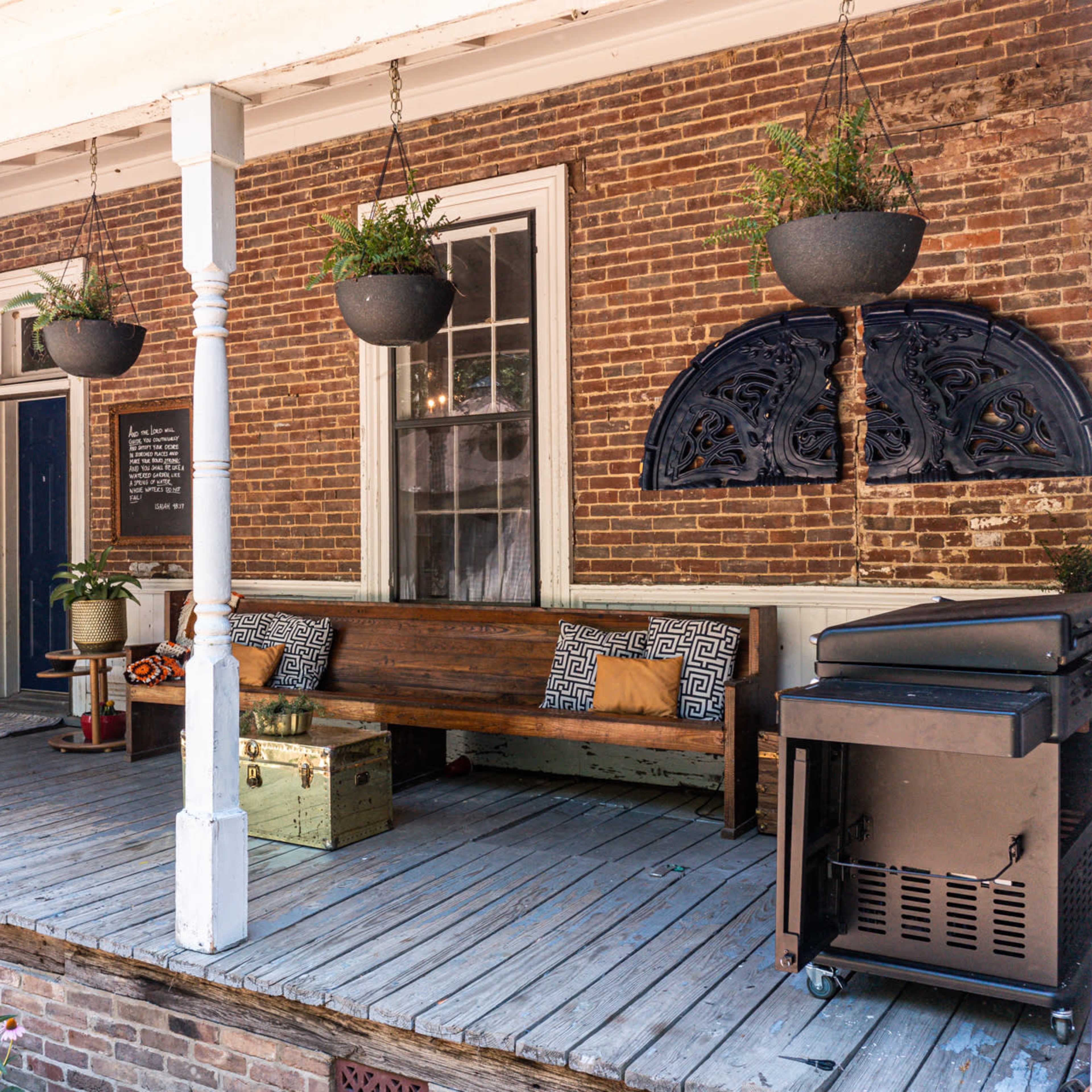 A rustic porch with a wooden bench, hanging planters, and a grill, adjacent to a brick wall and a white structure.