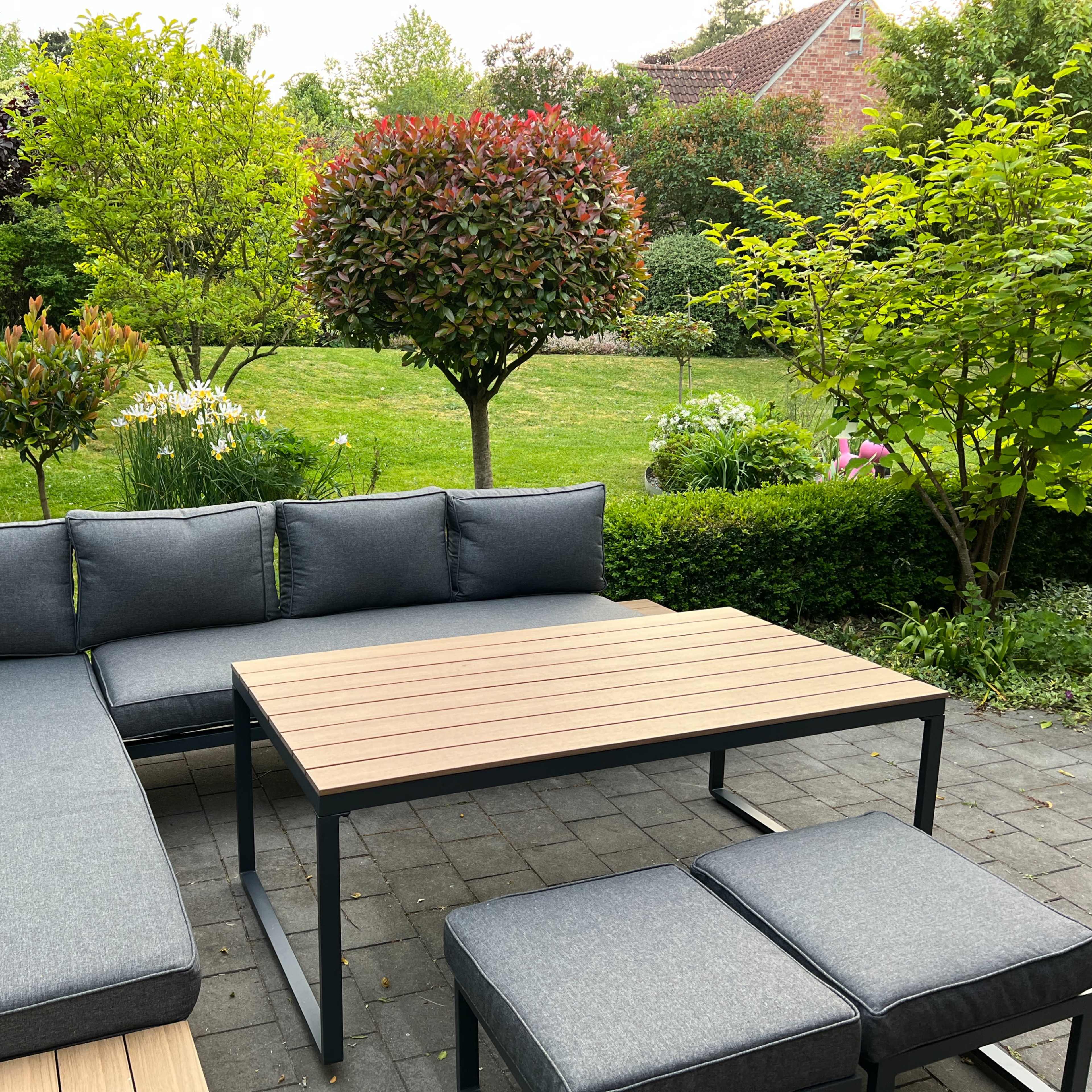 A modern outdoor seating area features a sectional grey couch and a wooden table surrounded by lush greenery and flowering plants.