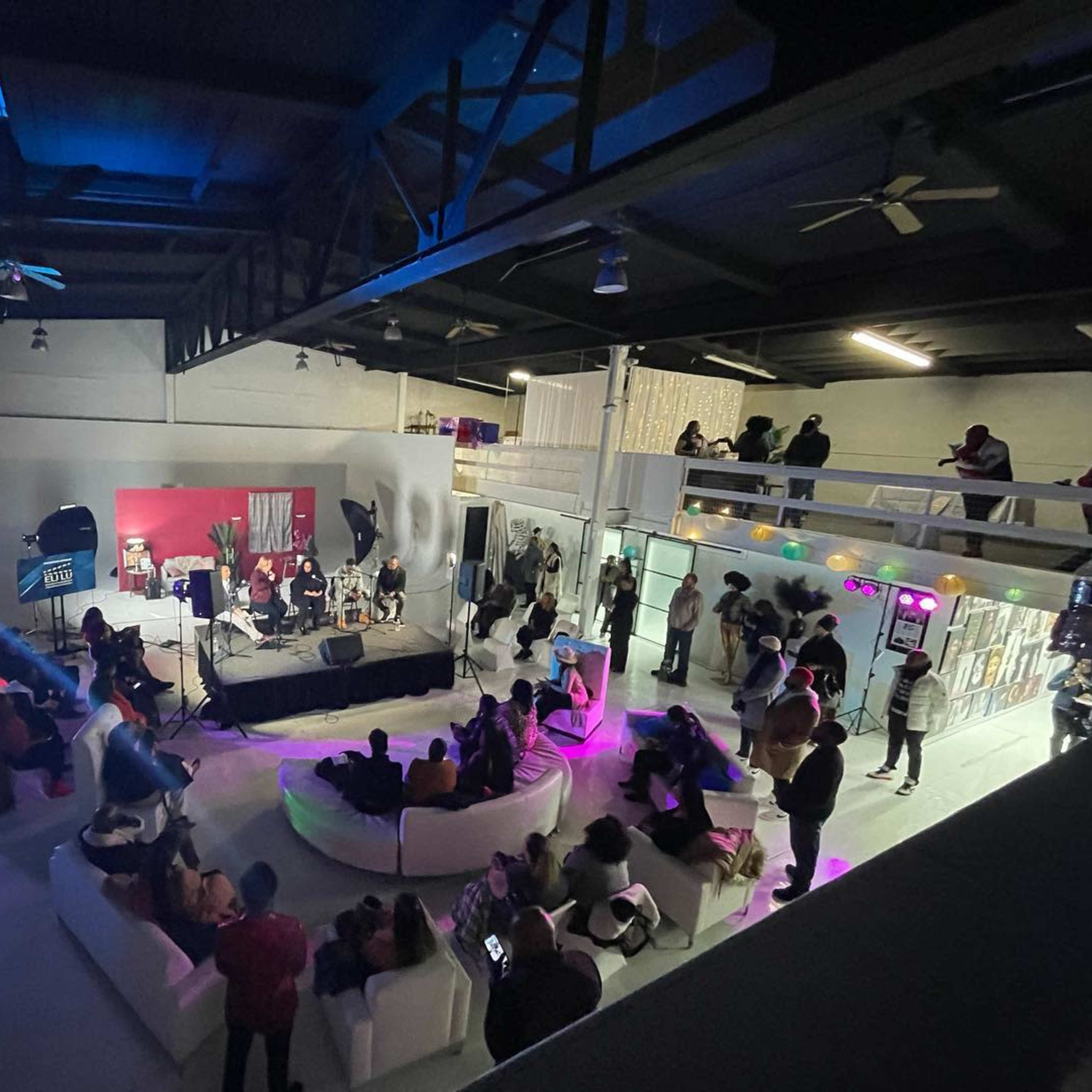 A diverse audience watches a live performance in a spacious, modern venue with elevated seating and various light setups.