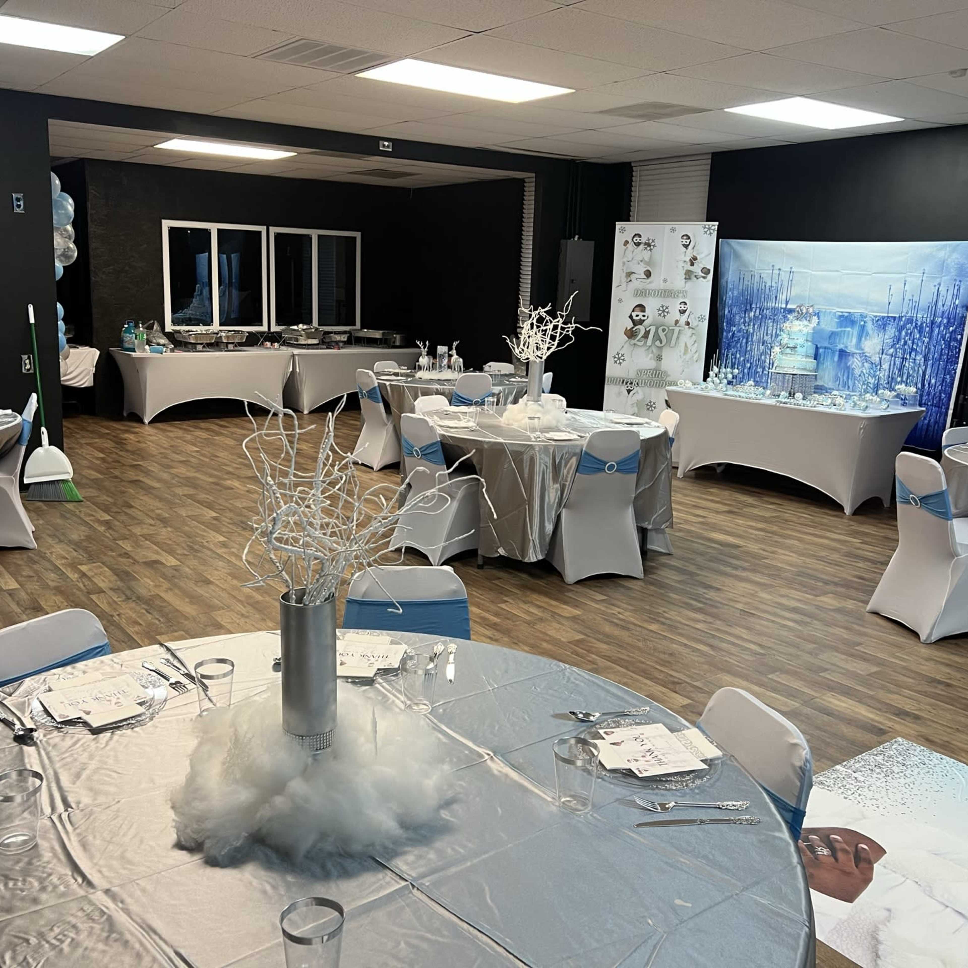 The room is set up for an event, featuring several round tables with silver tablecloths, chairs, and decorations, along with a display area in the background.