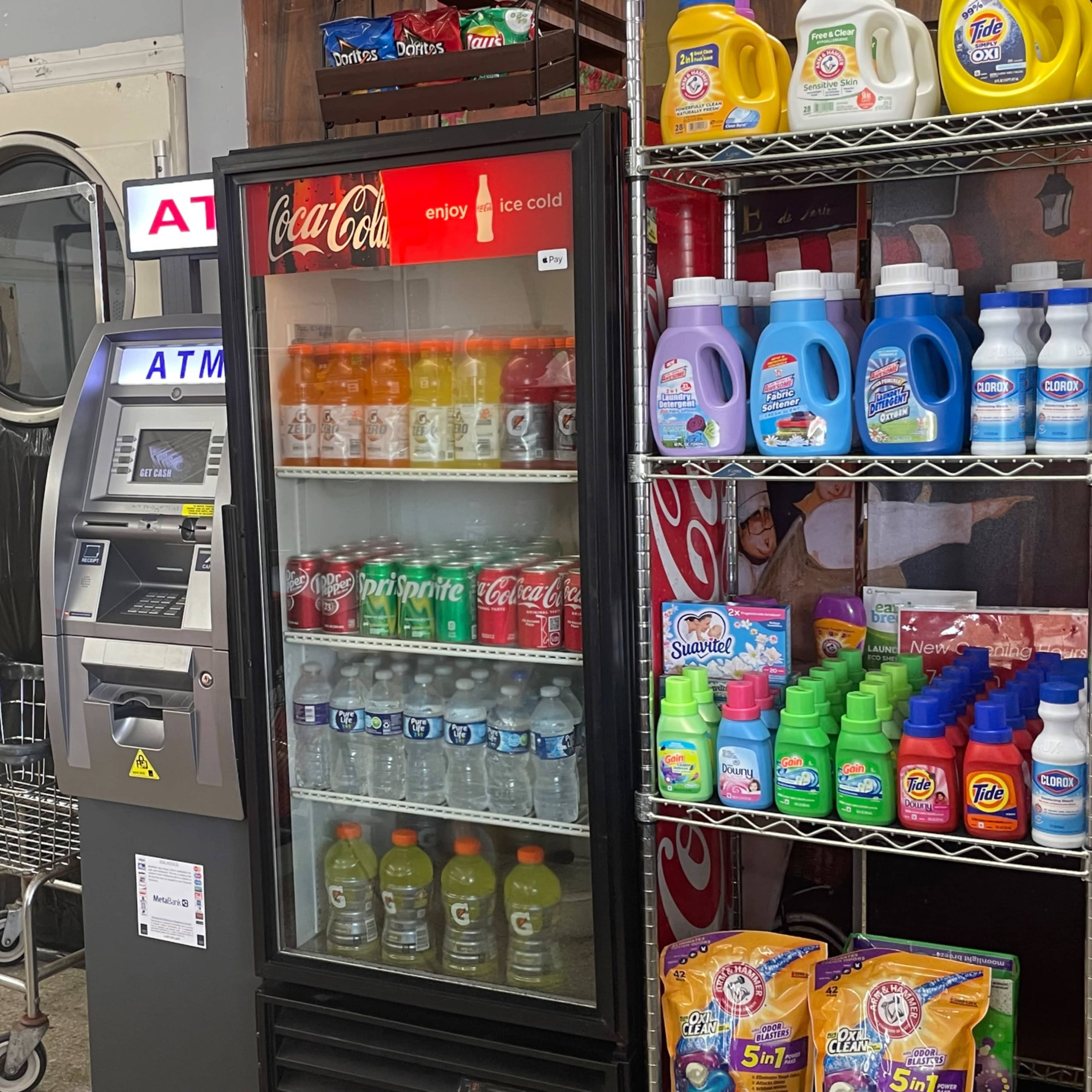 A laundromat with an ATM, a vending machine filled with drinks, and shelves stocked with laundry detergents and other household products.