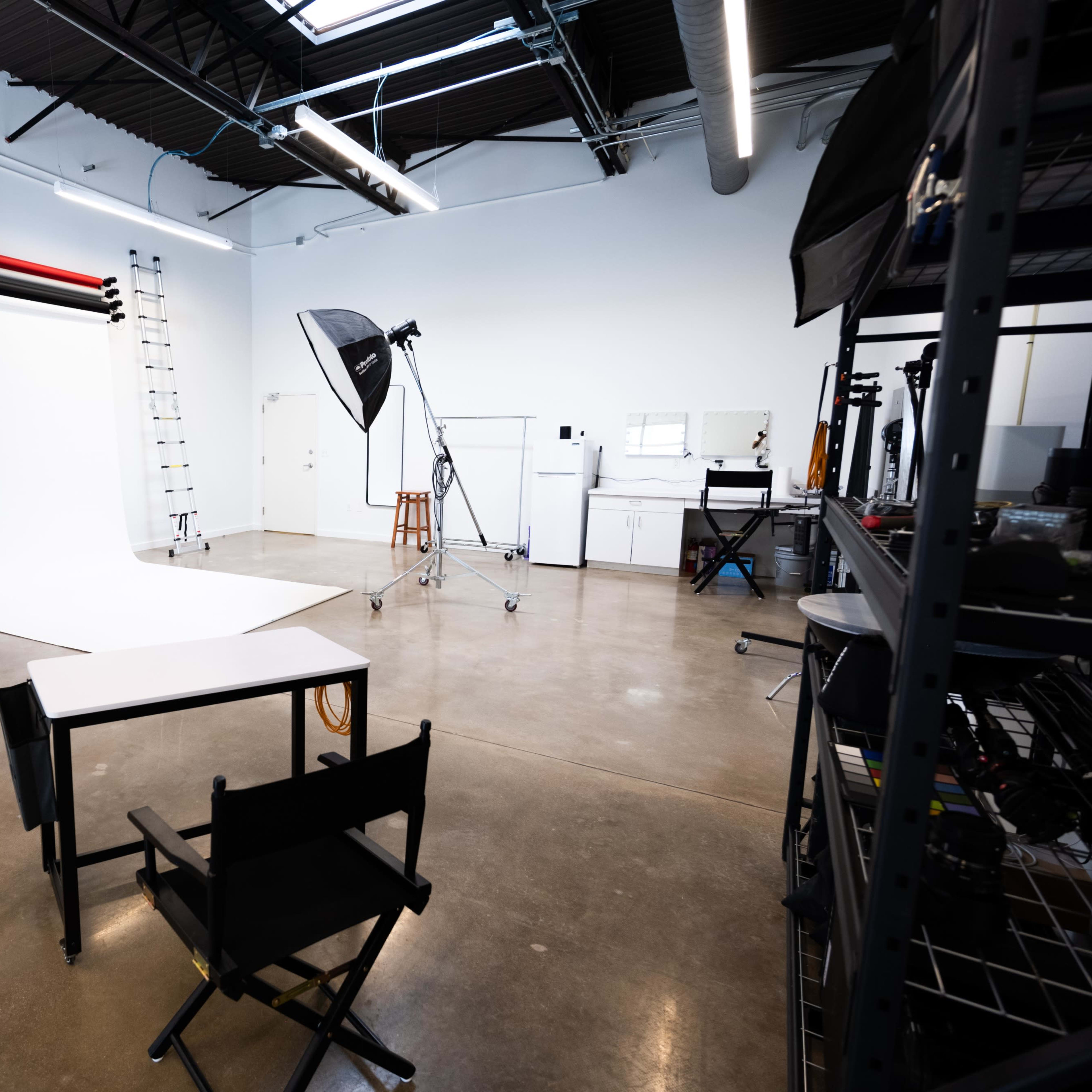 A photography studio with a white backdrop, lighting equipment, a ladder, and a table with a director's chair.