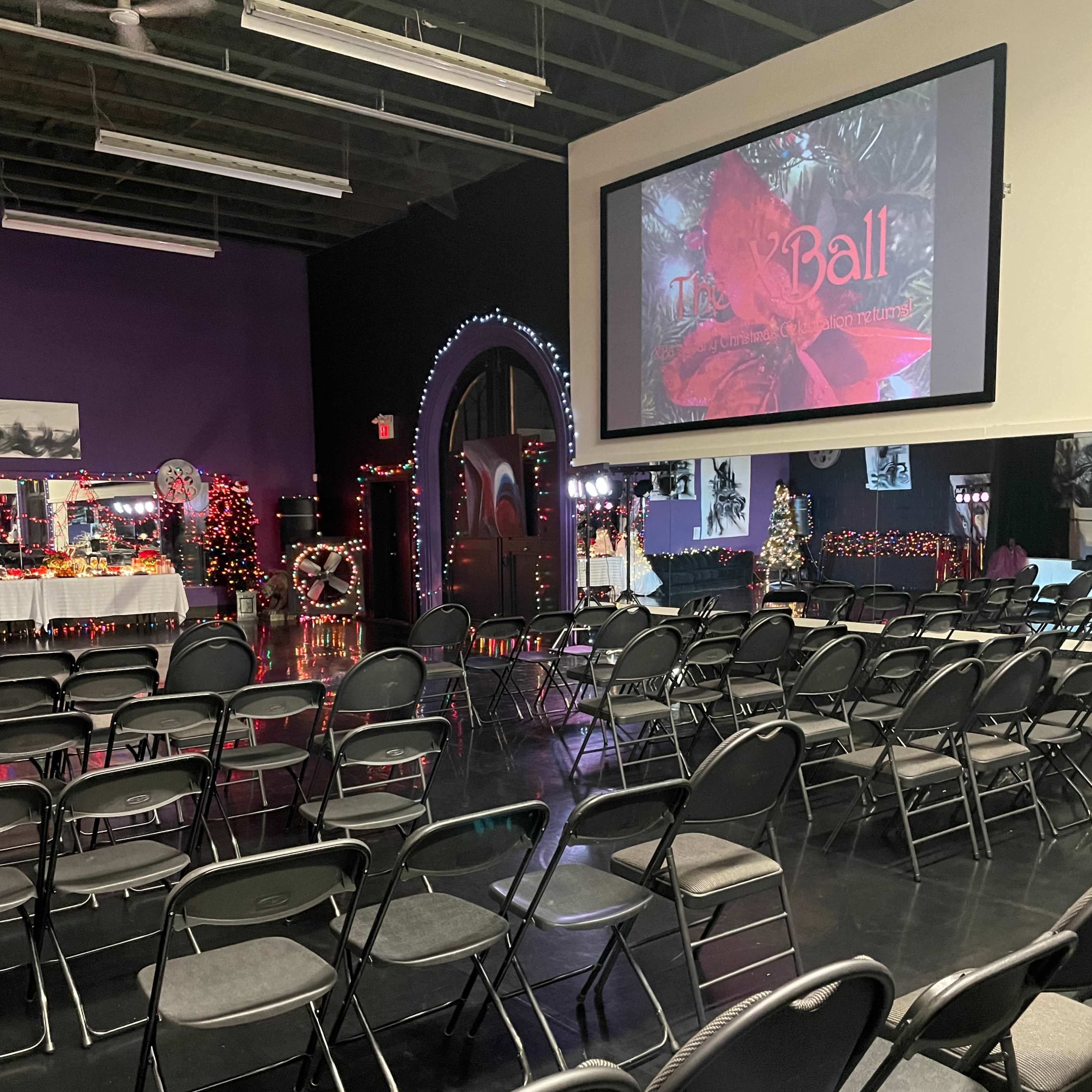 A hall setup for an event features rows of empty chairs facing a large screen displaying a red-themed graphic.