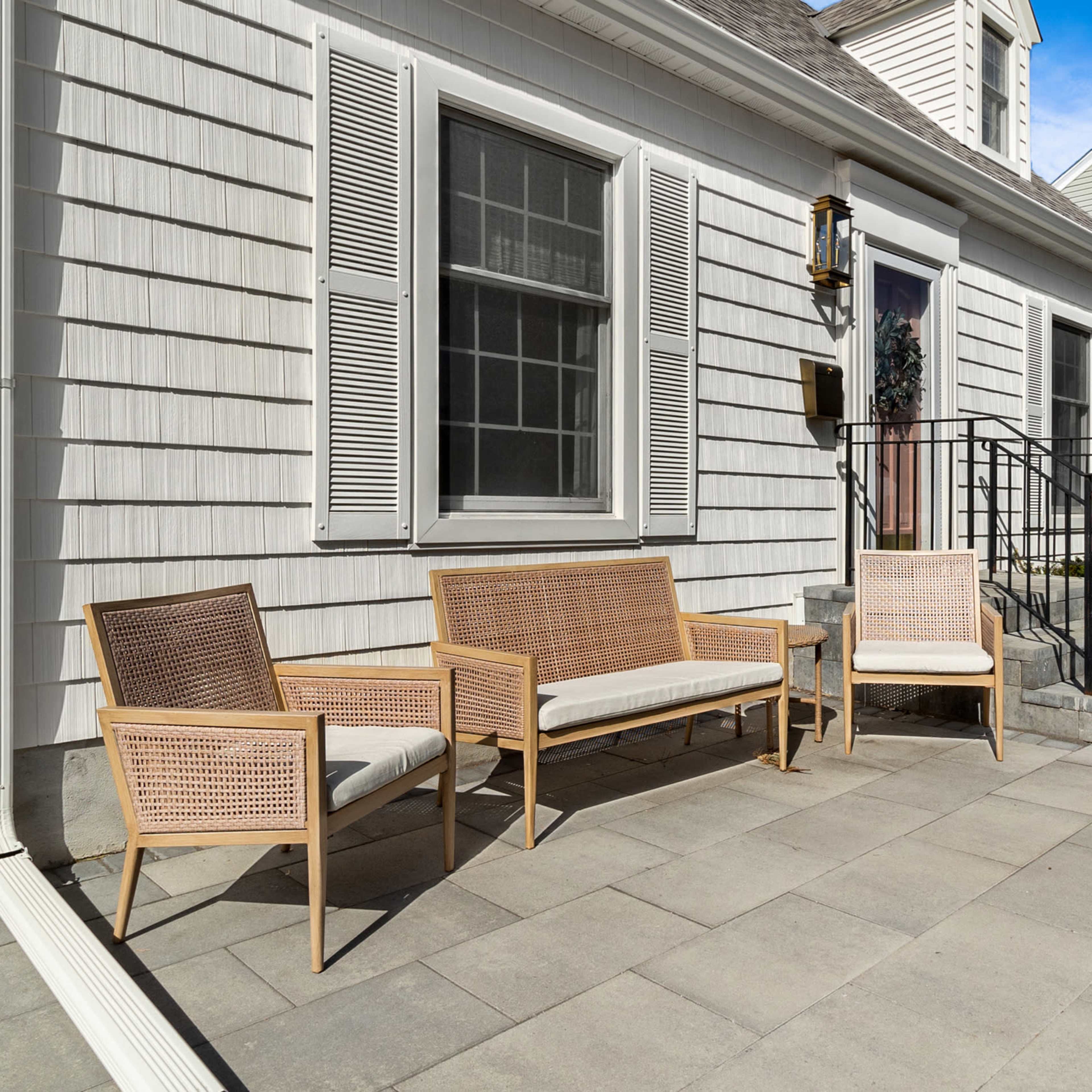 A front patio with a set of wicker chairs and a small table beside a house, featuring a stone pathway and steps leading to the entrance.