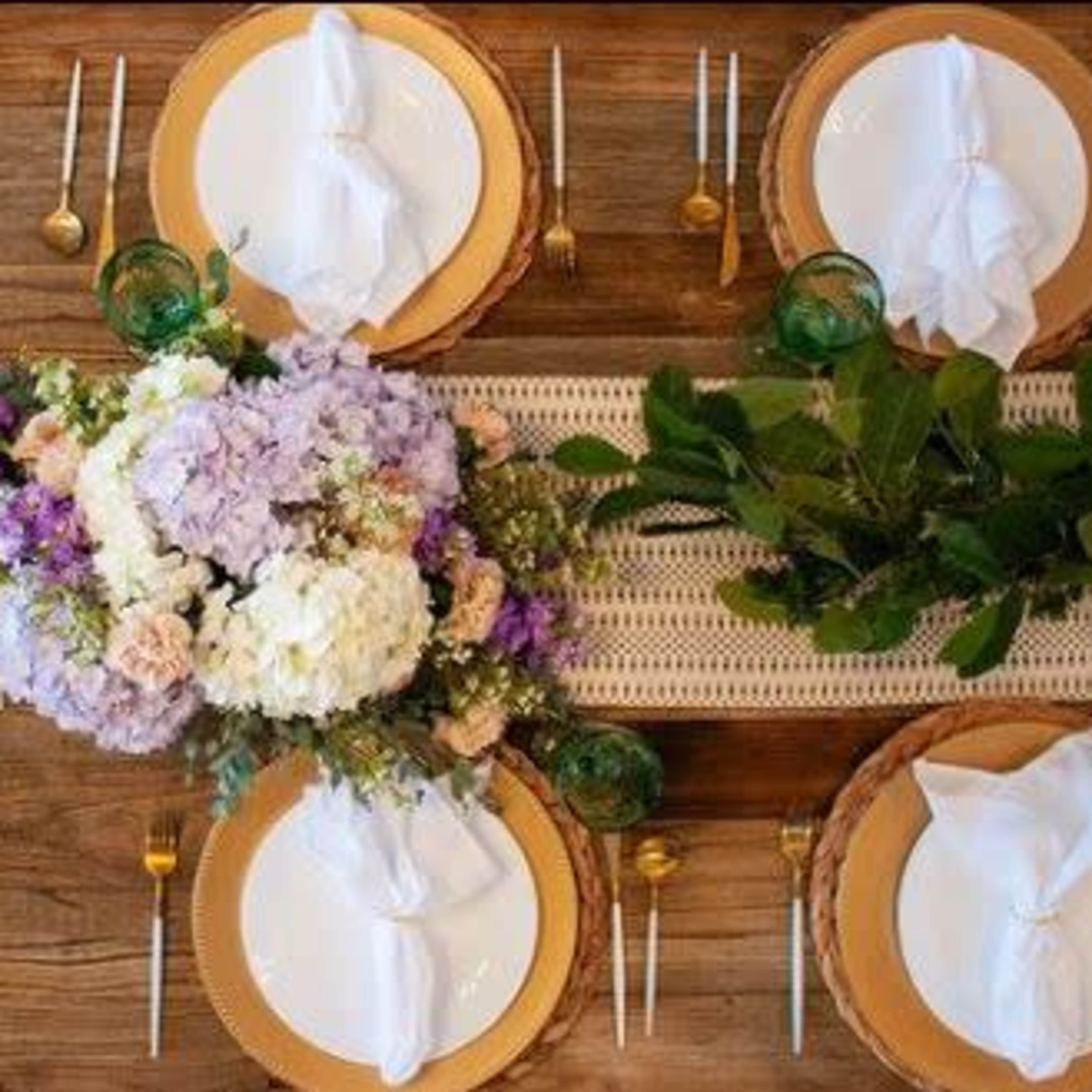 A decorated dining table is set with white plates, gold utensils, and a floral centerpiece, surrounded by greenery.
