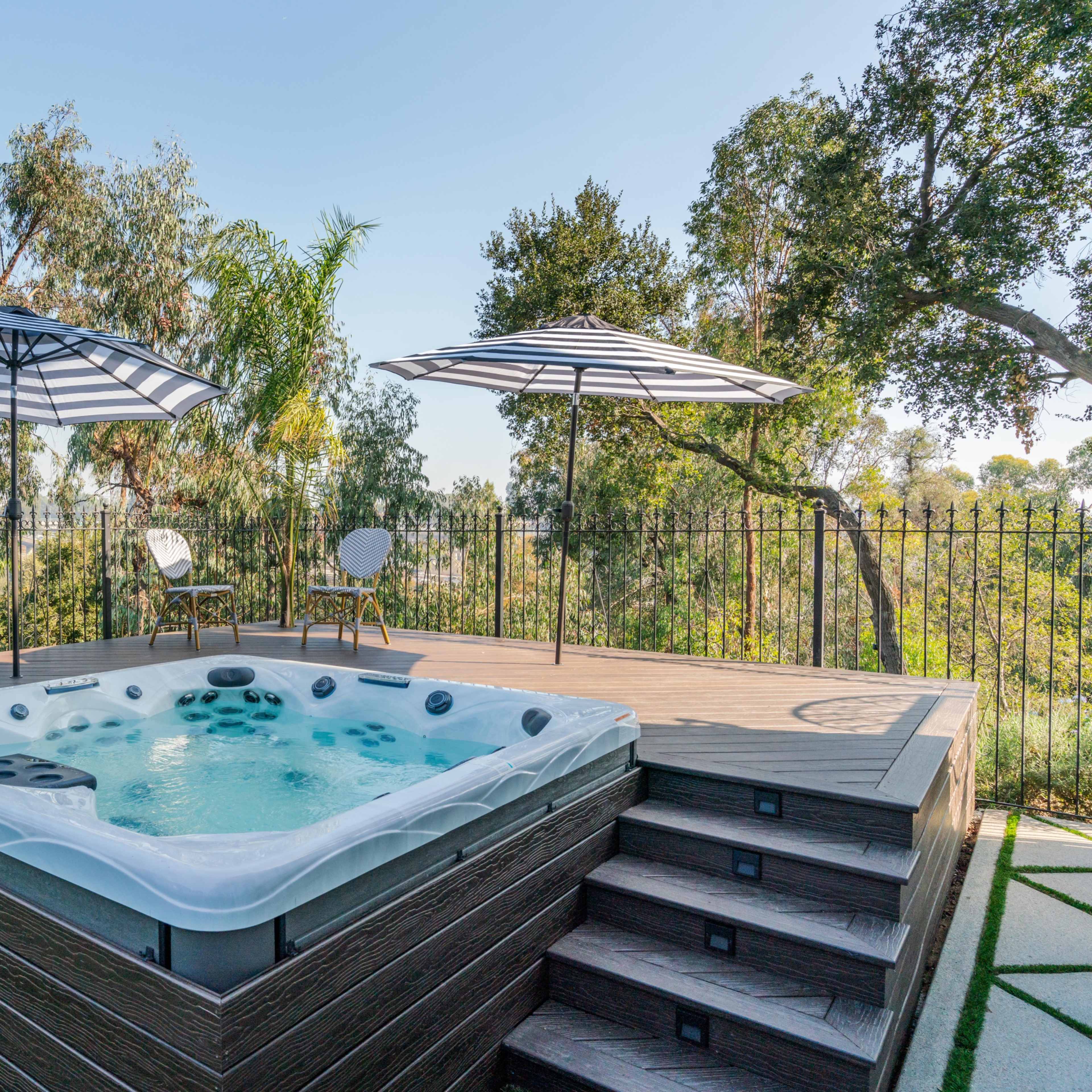 A hot tub on a wooden deck surrounded by greenery, with two umbrellas and a chair nearby.