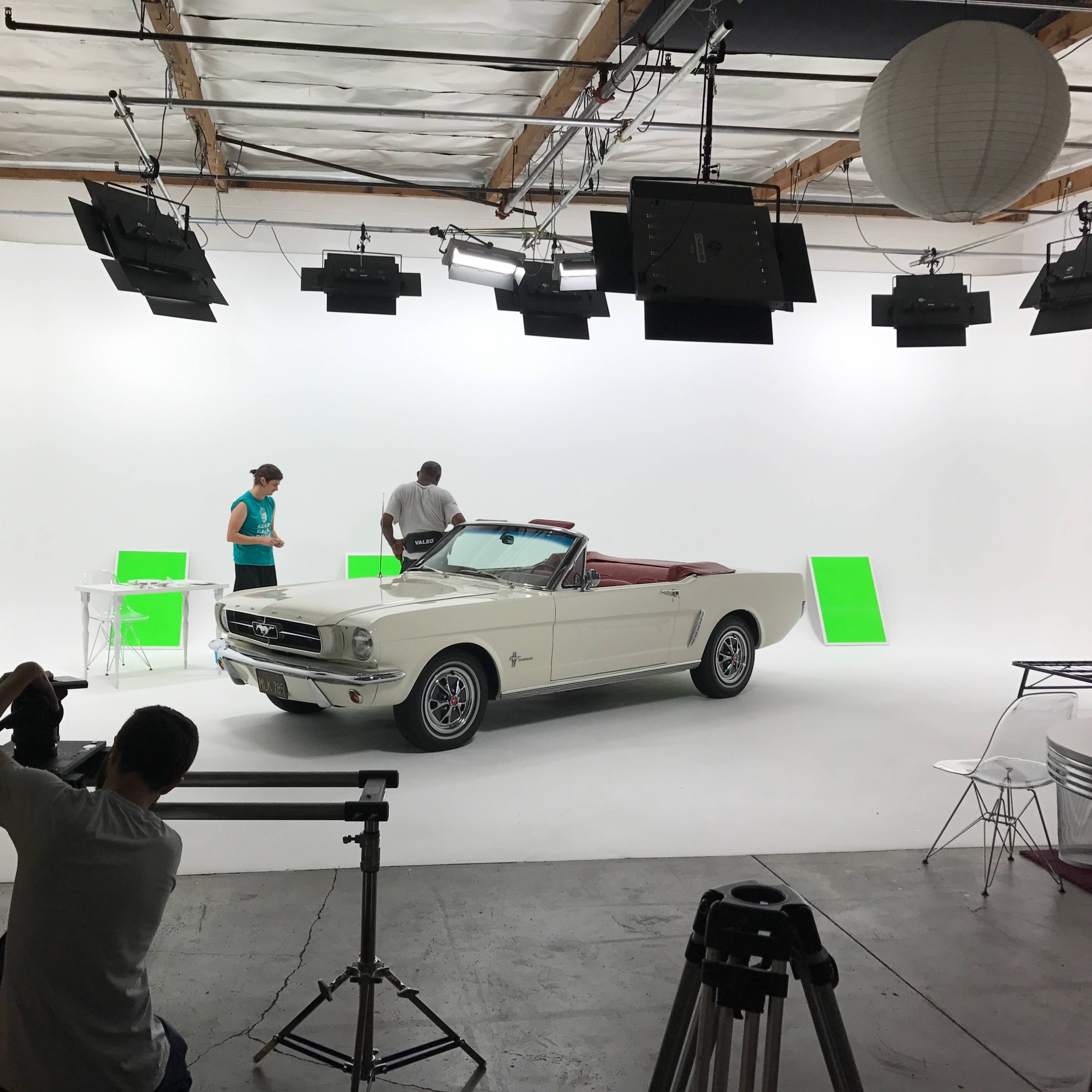 A vintage convertible car is displayed in a studio setting with two people preparing for a photoshoot among various lighting equipment and green screens.