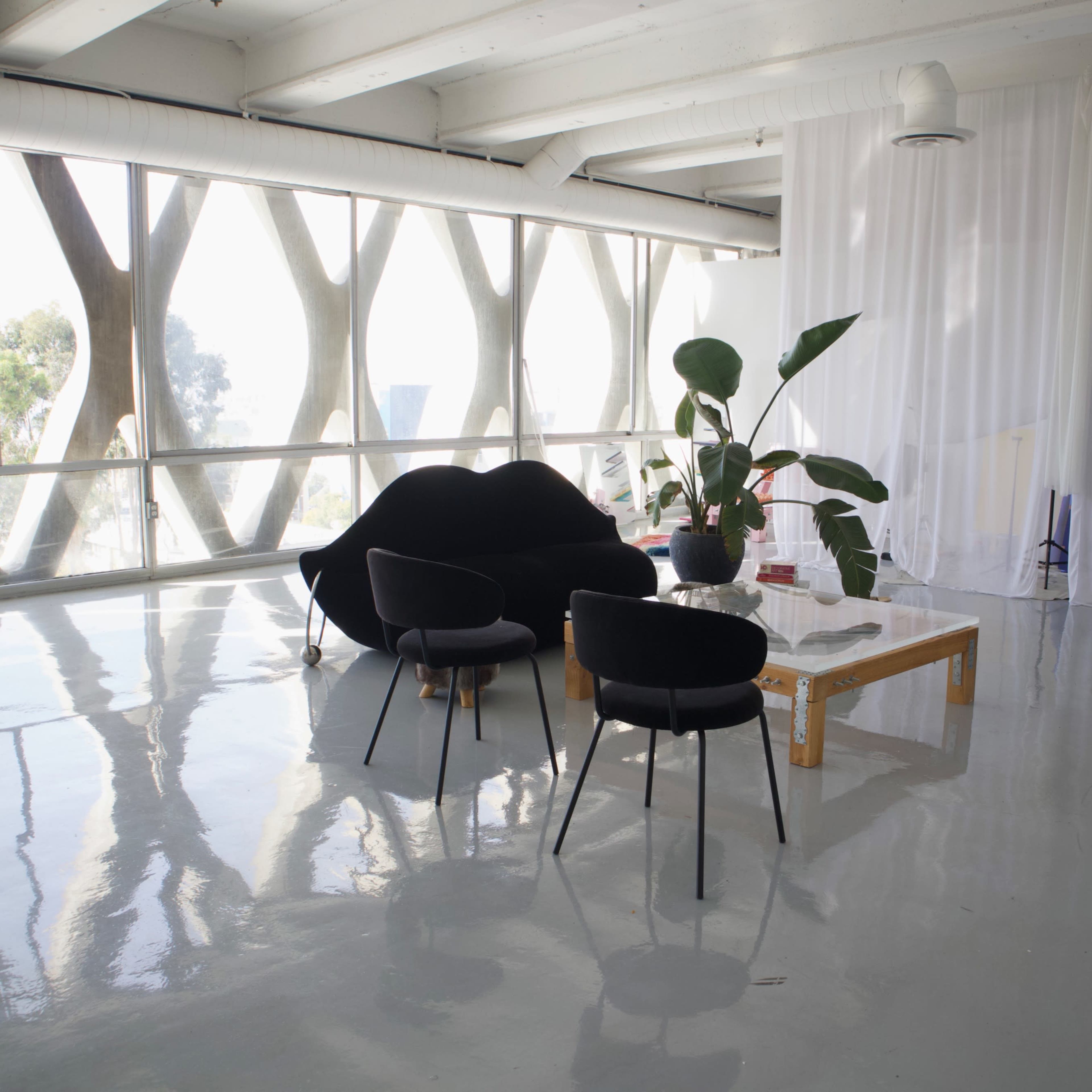 A modern interior space features large windows, a black sofa, two black chairs, a wooden coffee table, and a potted plant.