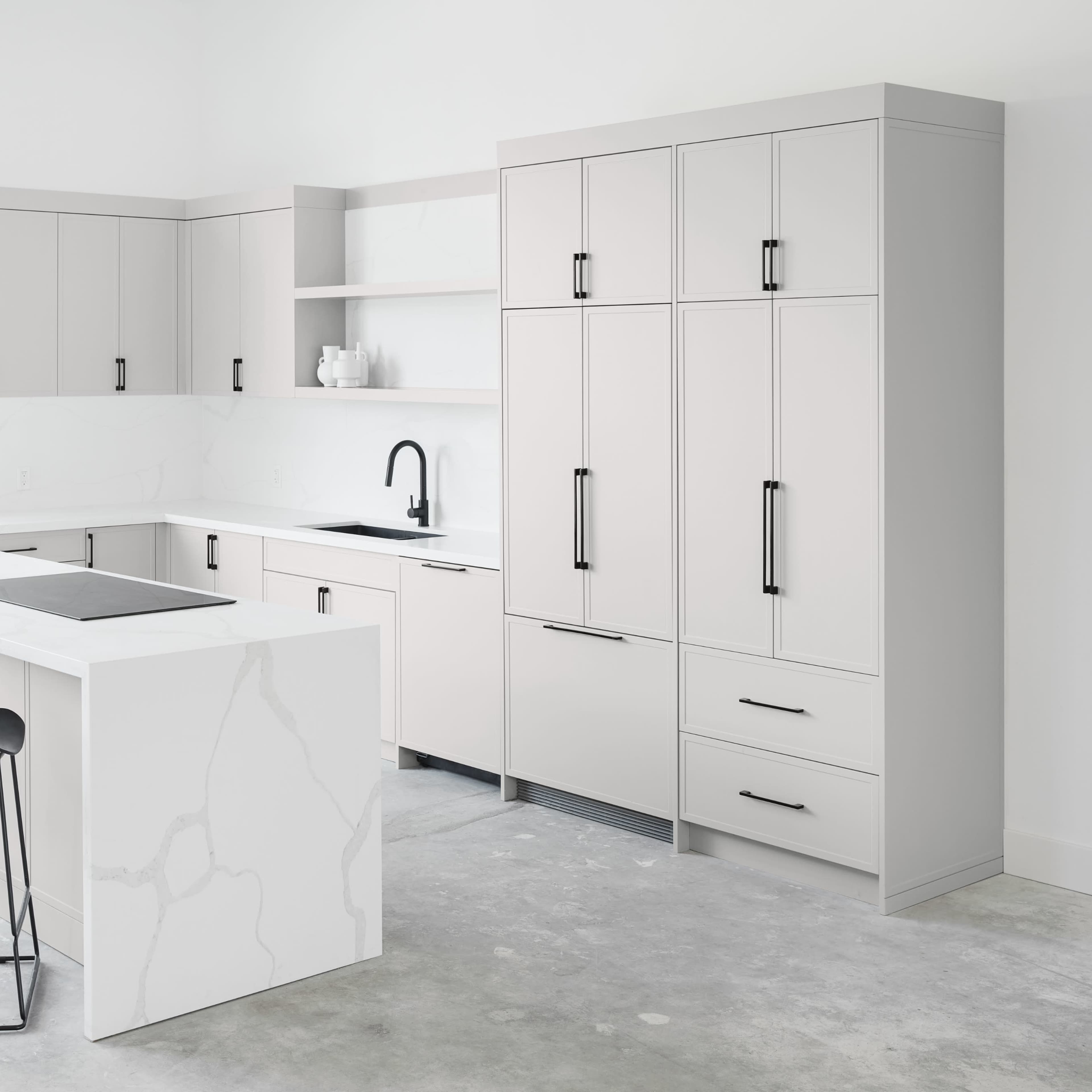 A modern kitchen features minimalist cabinetry, a large island with seating, and sleek appliances against a light-colored background.