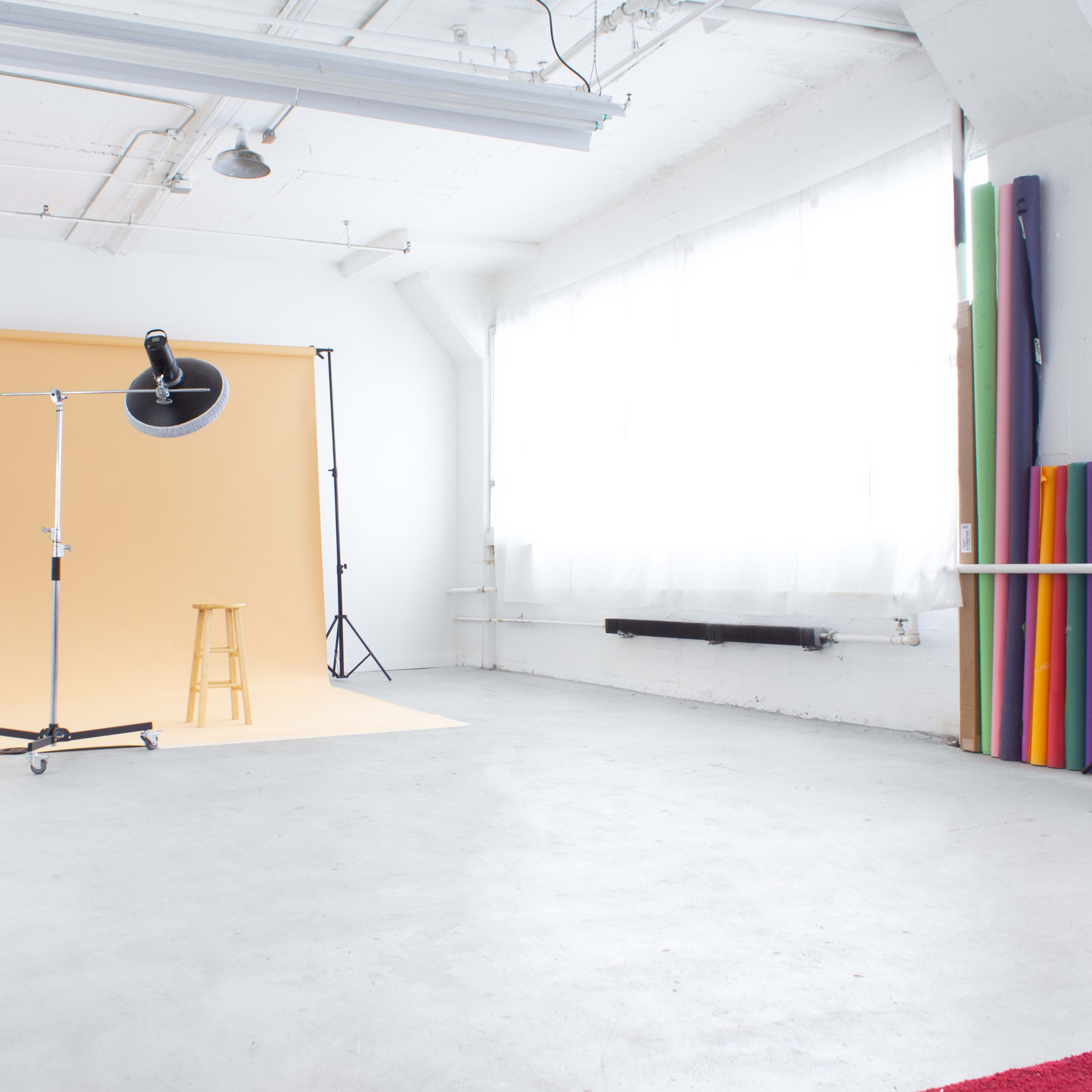 A well-lit photo studio featuring a beige backdrop, a wooden stool, and colorful backdrops stored against the wall.