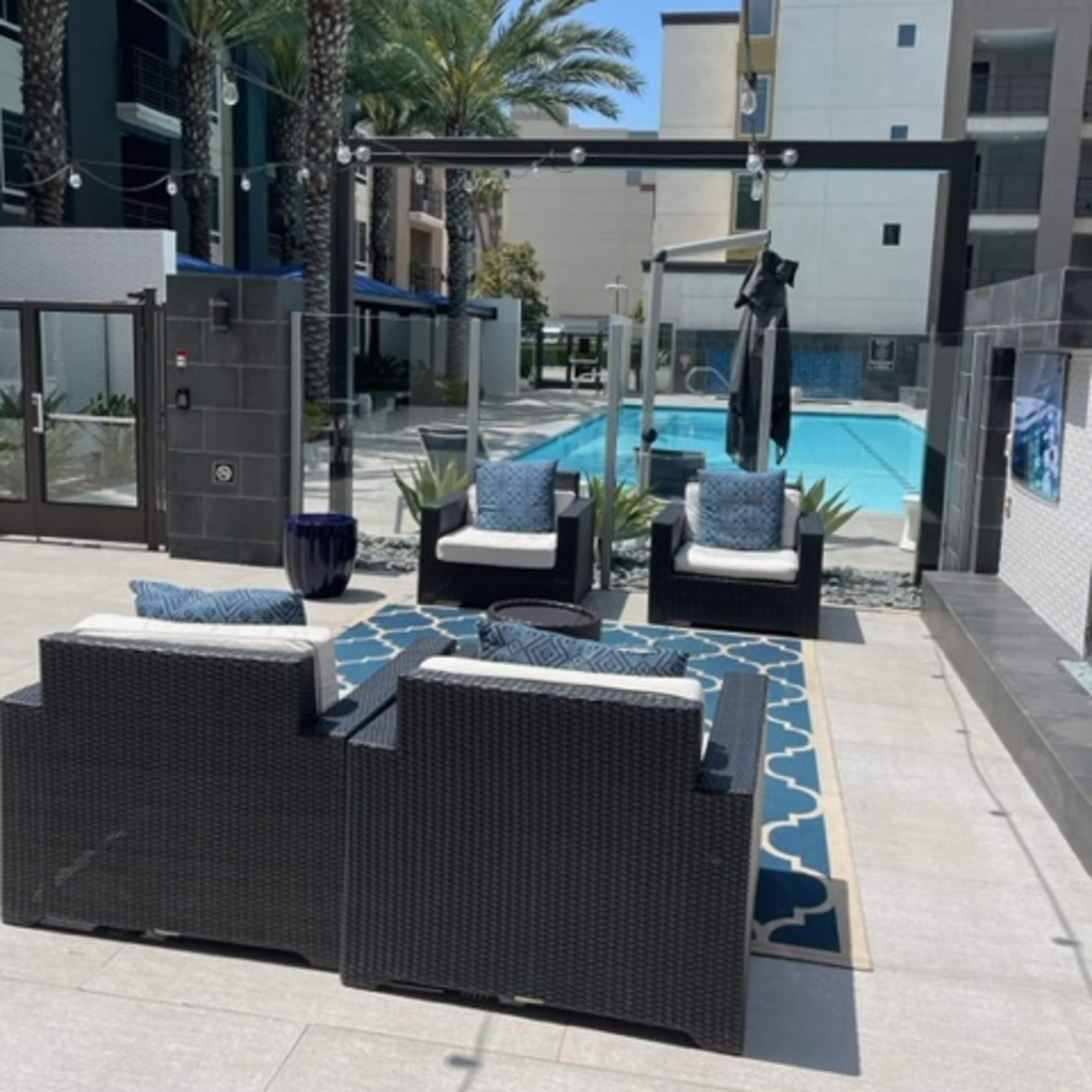 The scene shows a modern outdoor lounge area with four wicker chairs arranged on a patio beside a swimming pool surrounded by palm trees.